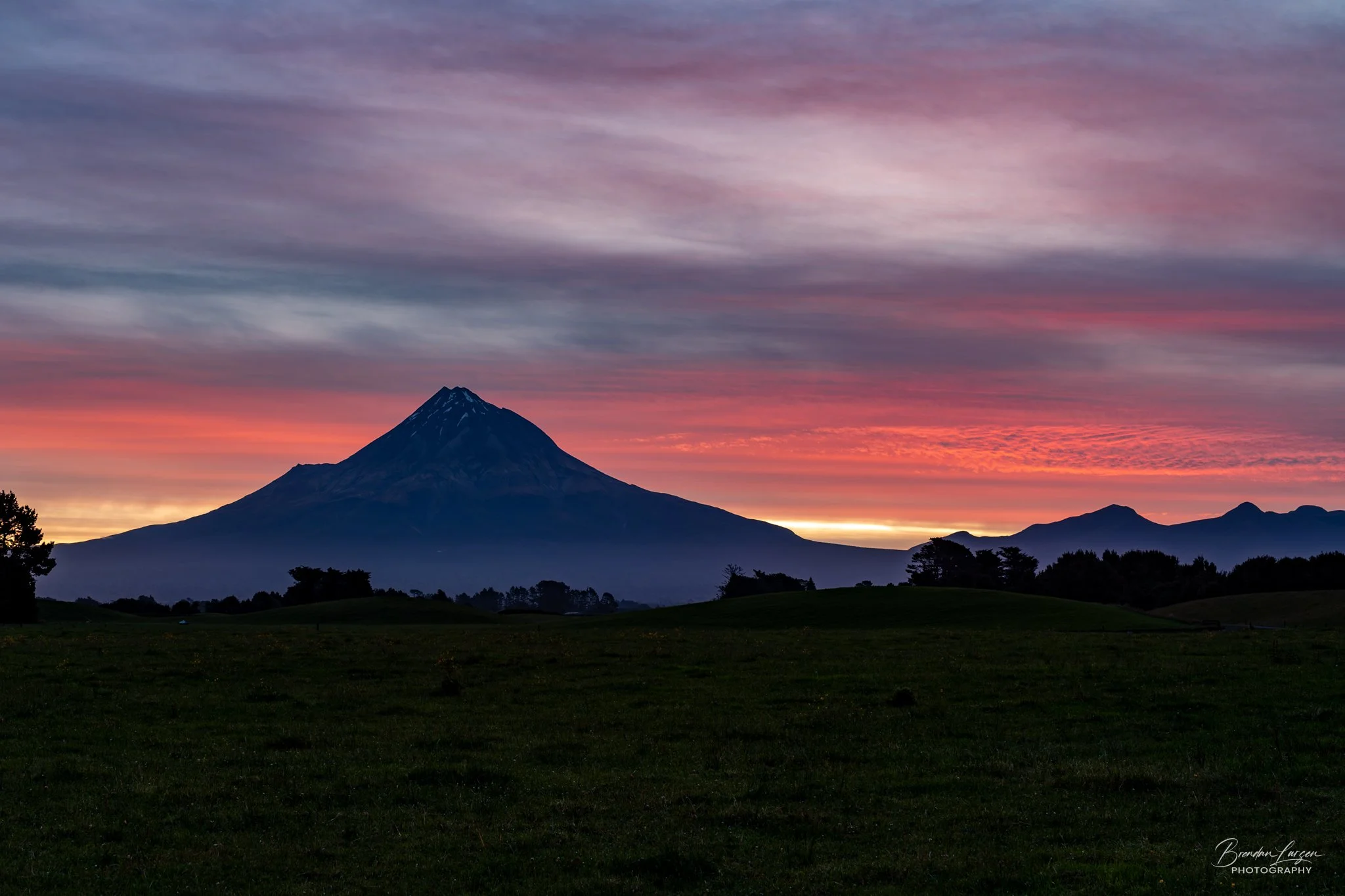 A mountain with a snow-capped peak against a colorful sunset sky with shades of pink, purple, orange, and blue, and a grassy field in the foreground.