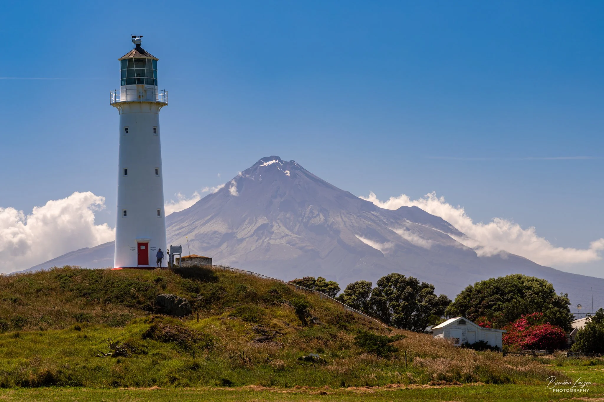 A lighthouse on a grassy hill with a person standing near it, and Mount Taranaki in the background under a blue sky with some clouds.