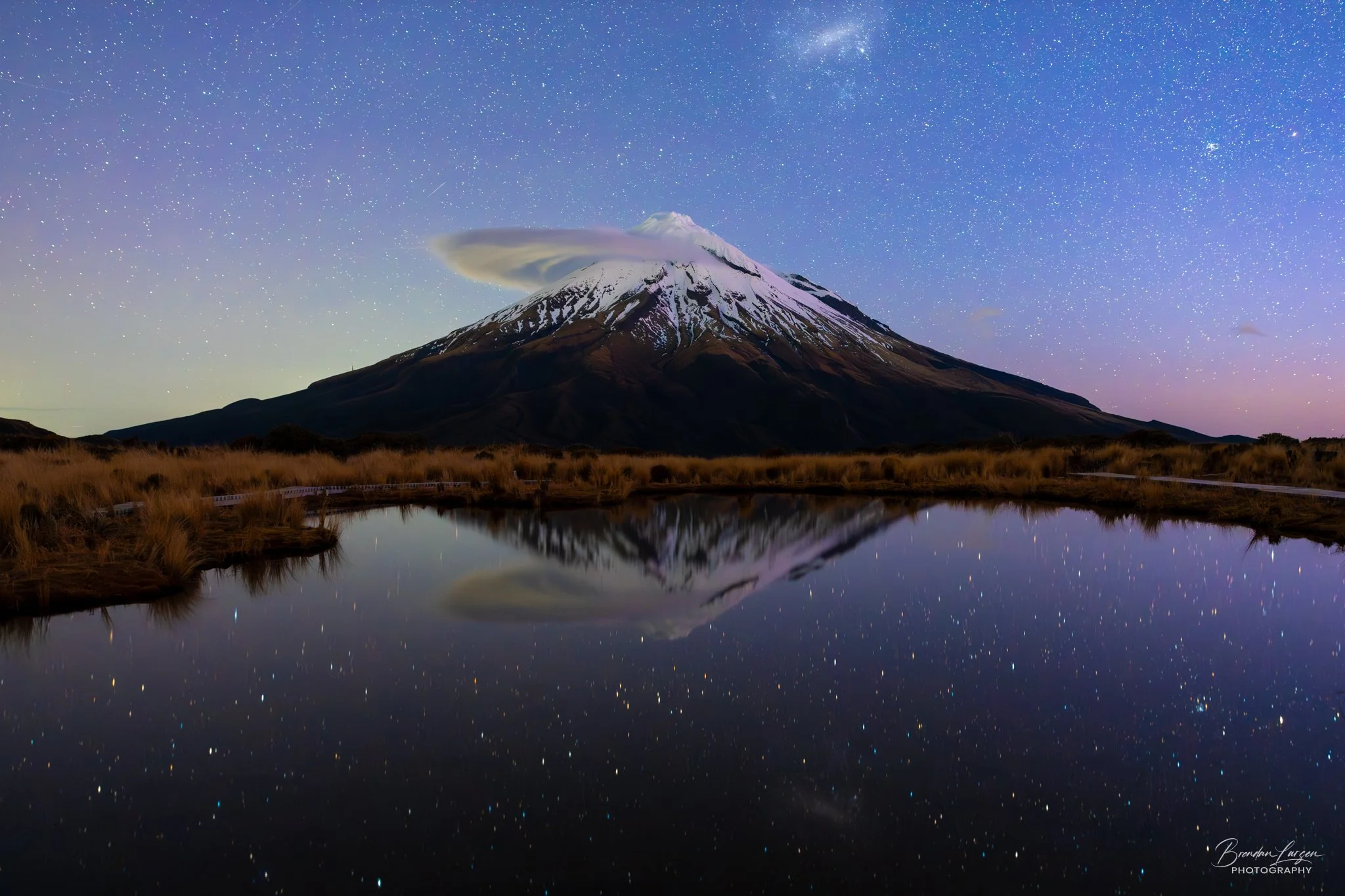 Starry night sky above Mount Taranaki with snow-capped peak, reflected in a calm body of water surrounded by grassy terrain.