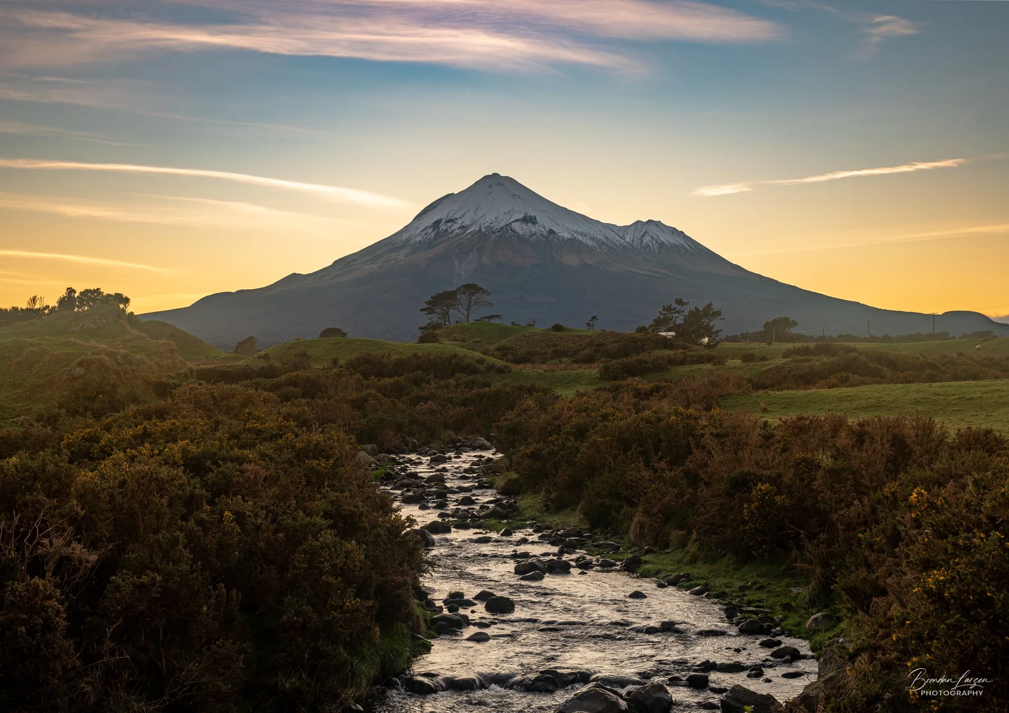 A mountain with snow on its peak against a sunset sky, with a flowing river and green vegetation in the foreground.
