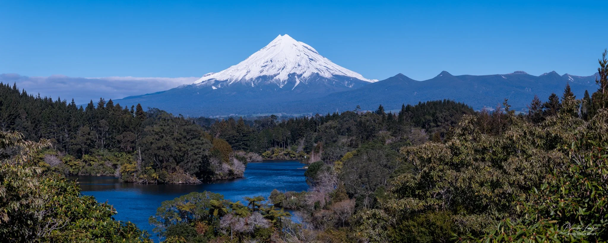 Snow-capped mountain in the background with a forest and river in the foreground on a clear day.