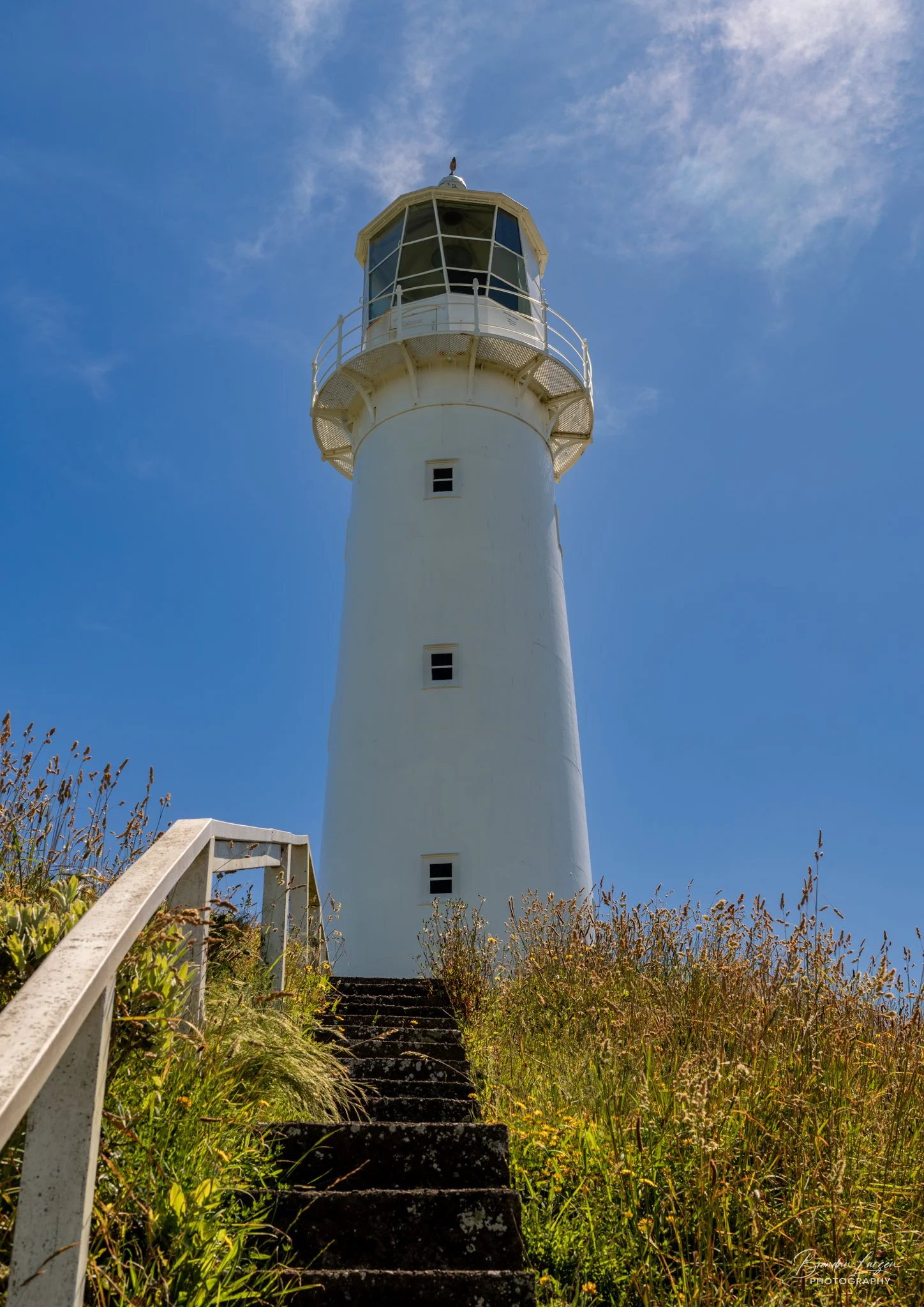 White lighthouse on a grassy hillside with steps leading up to it, set against a bright blue sky.
