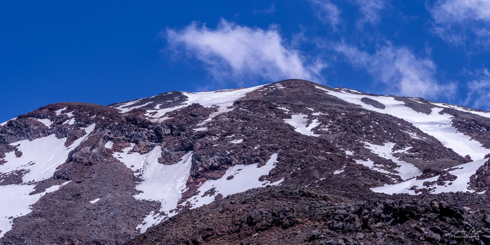 Snow-covered mountain with dark rocky terrain and a blue sky with a small white cloud