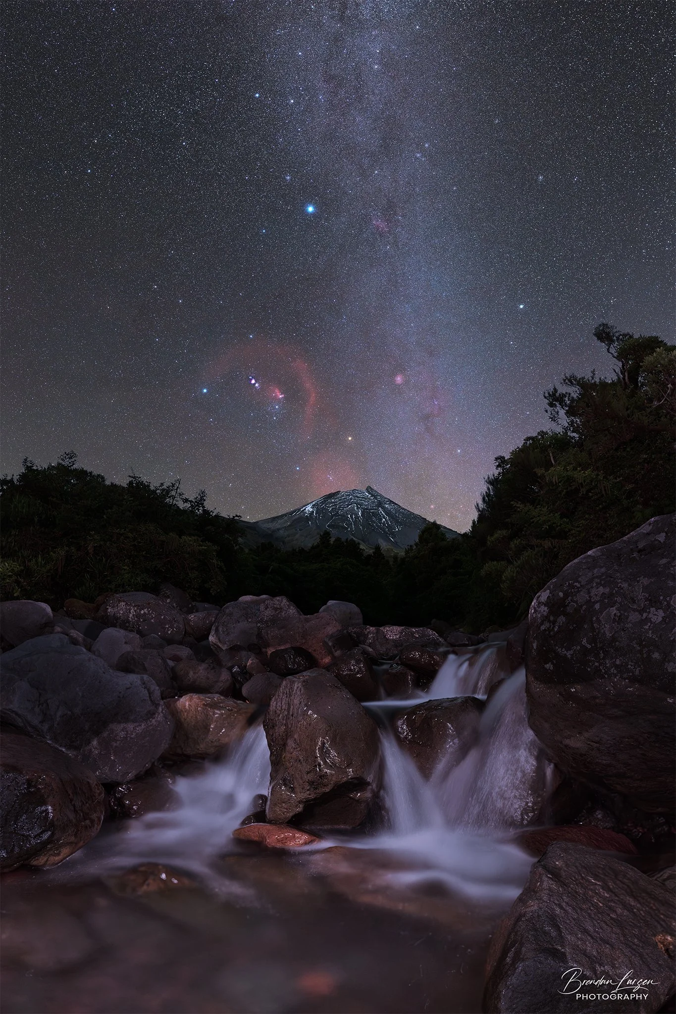 Nighttime landscape with a starry sky, mountain, forest, and a flowing stream in the foreground.
