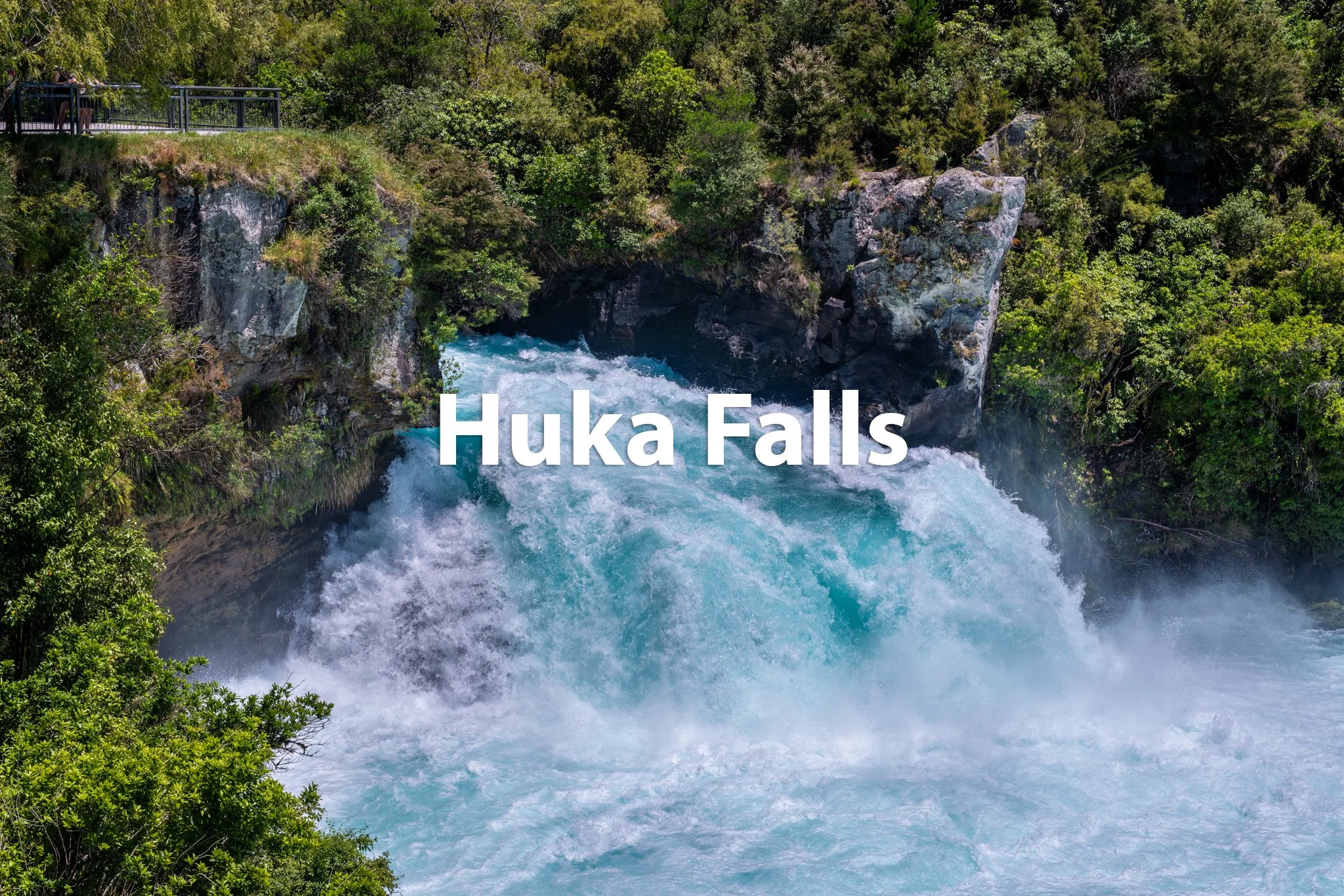 Huka Falls with rushing water surrounded by lush green foliage and rocks, and a walkway with a railing at the top left.