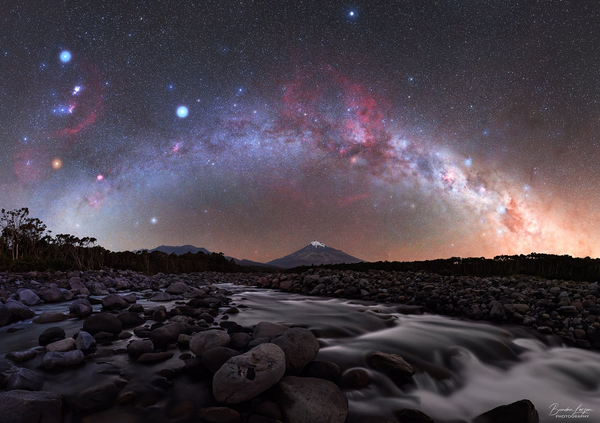 Night sky filled with stars and the Milky Way galaxy arching across the sky, with a snow-capped mountain and a rocky river in the foreground.