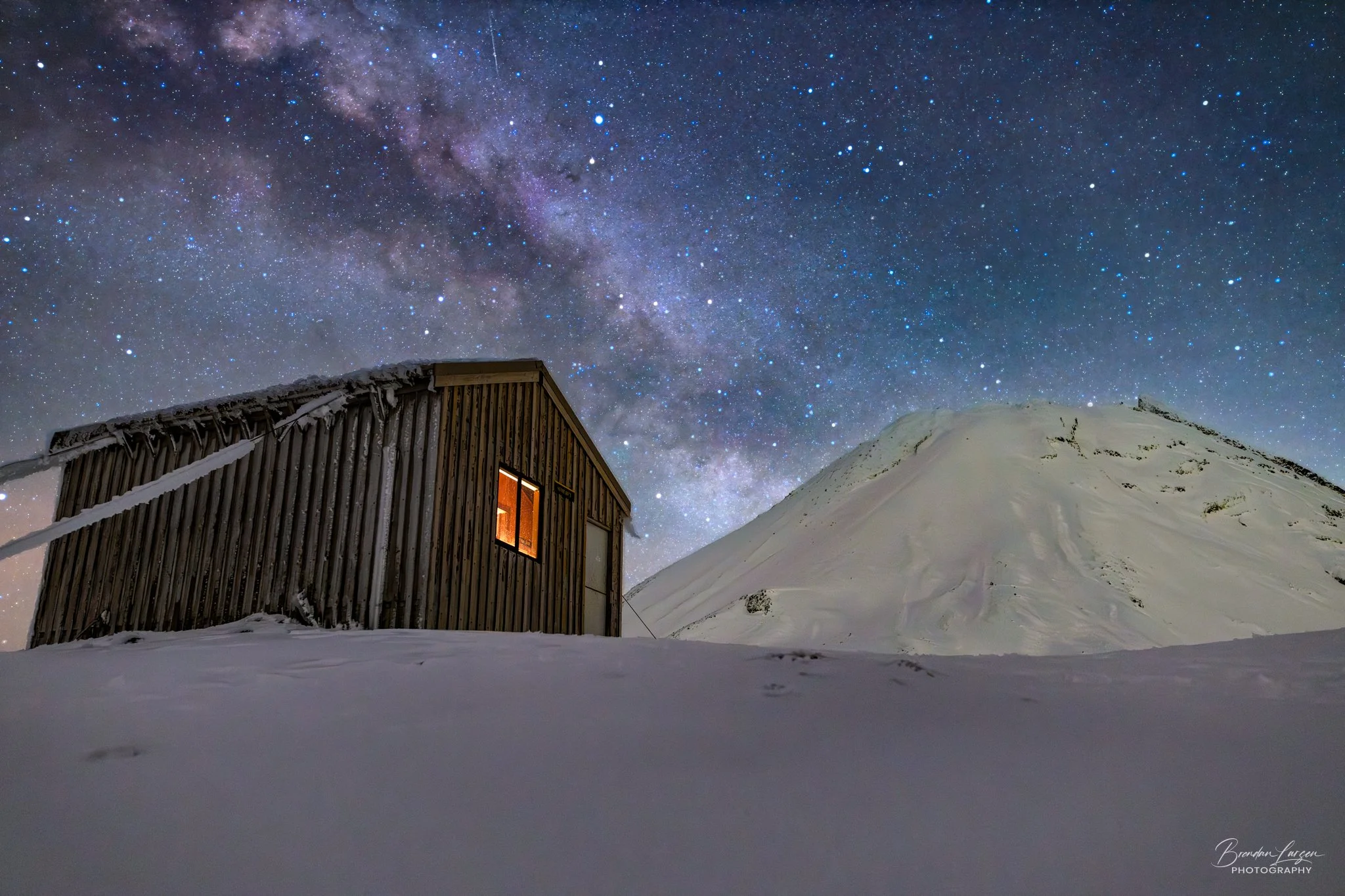 A wooden cabin with warm light inside, snow-covered ground, and a snow-covered mountain in the background under a starry night sky filled with the Milky Way galaxy.