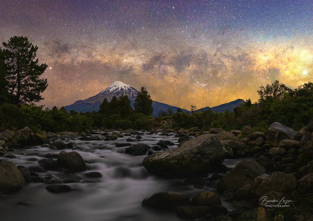 Night sky with stars and the Milky Way over a mountain with a snow-capped peak, a river flowing over rocks, surrounded by trees and greenery.