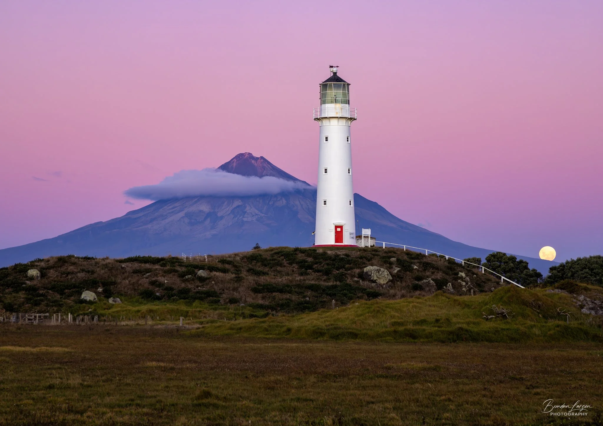 A white lighthouse on a grassy hill with a mountain in the background, pink sky, and the moon near the horizon.