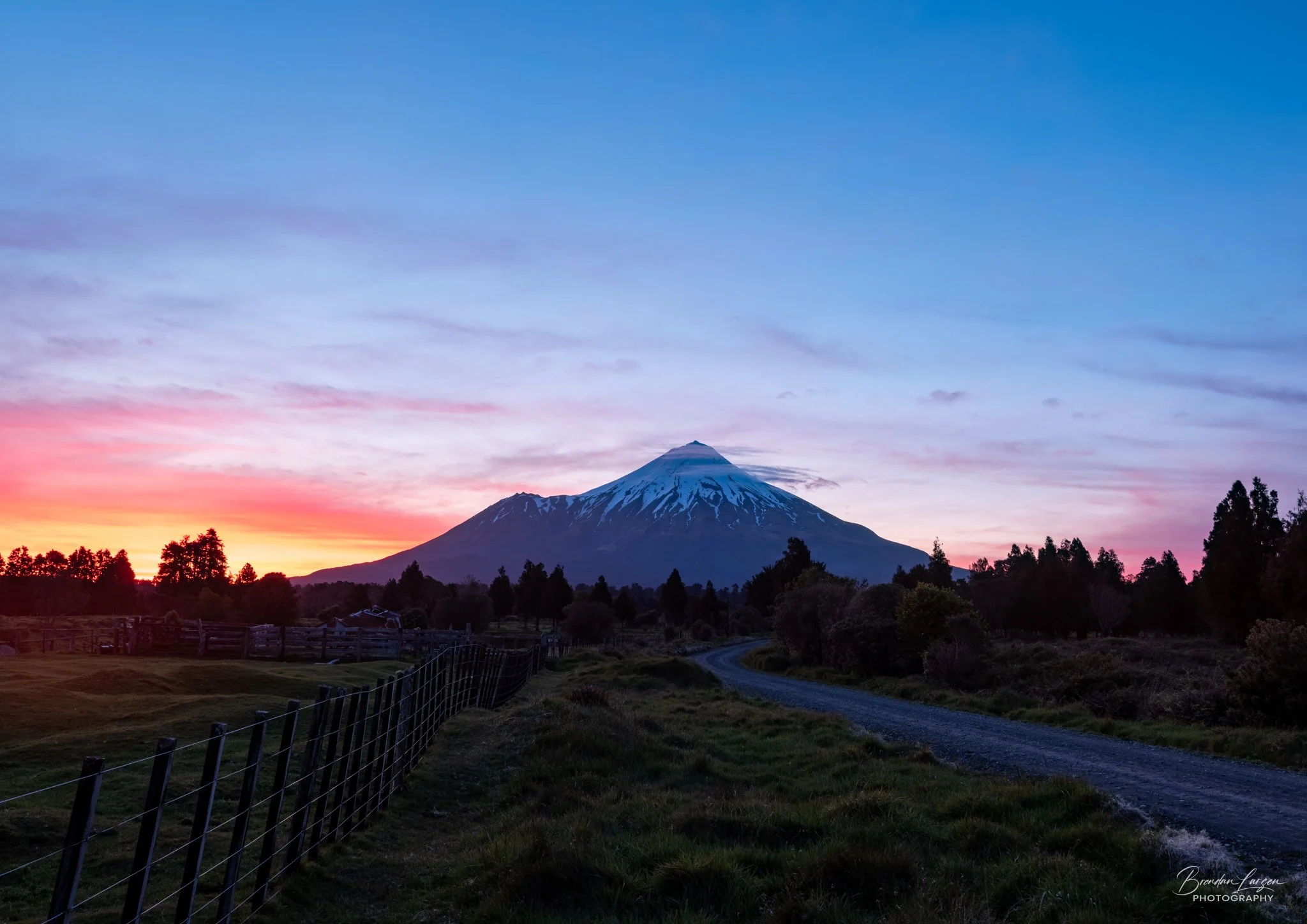 Scenic landscape of a snow-capped mountain at sunset with a dirt road and trees in the foreground.