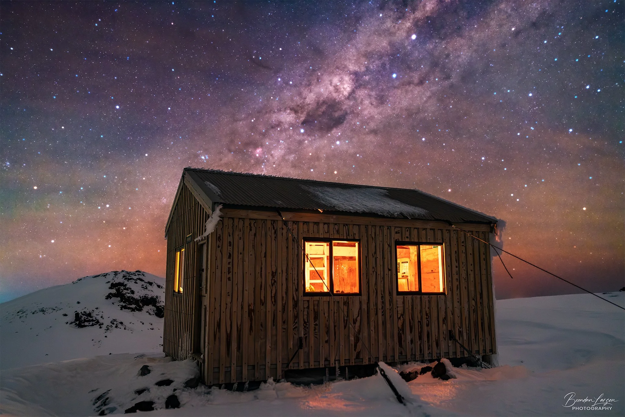 A wooden cabin with warm yellow-orange lights inside, standing in a snowy landscape under a night sky filled with stars and the Milky Way galaxy.