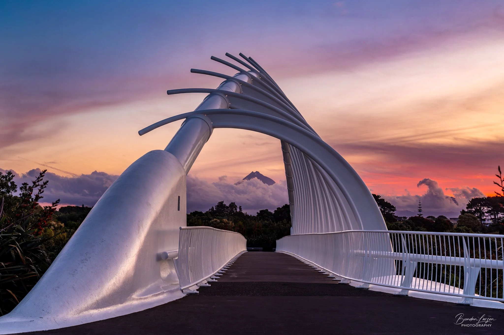 White modern bridge with curved arches and railings at sunset, with mountains and cloudy sky in the background.
