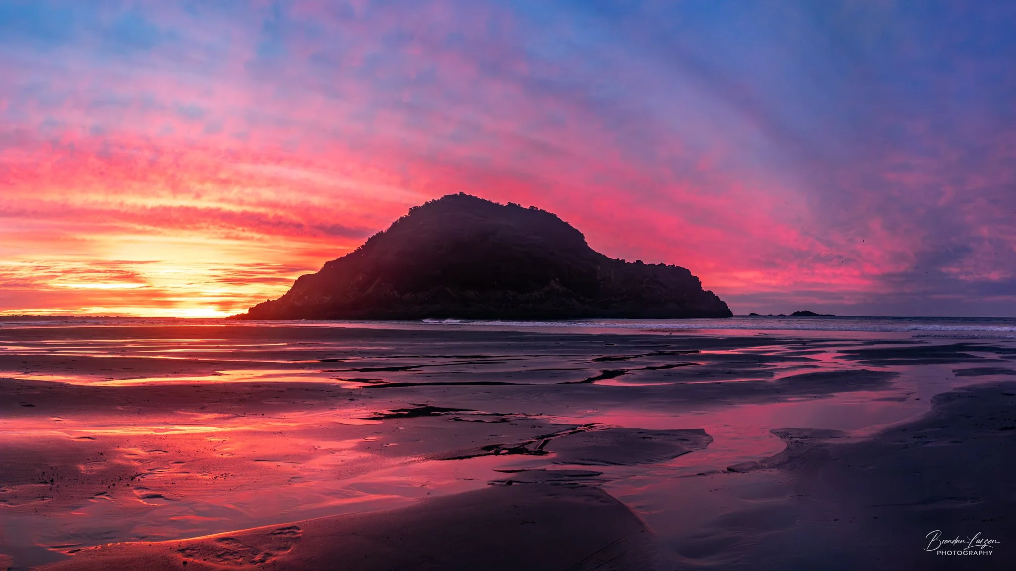 A landscape of a beach at sunset with pink and purple clouds in the sky, a rocky island in the distance, and calm water reflecting the vibrant colors.