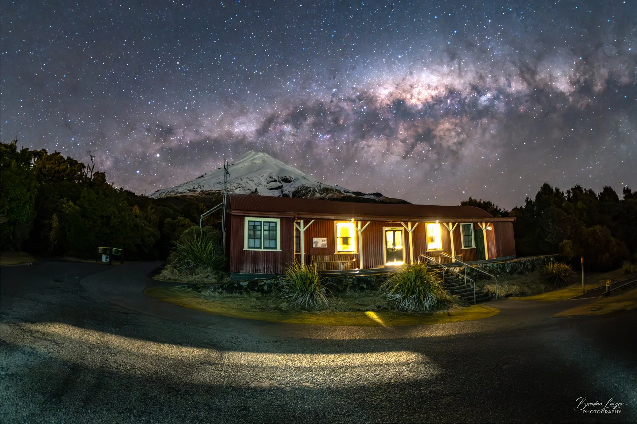 A small red building with lights on, situated in a rural area at night. In the background, a snow-capped mountain and a clear starry sky with the Milky Way galaxy visible overhead.
