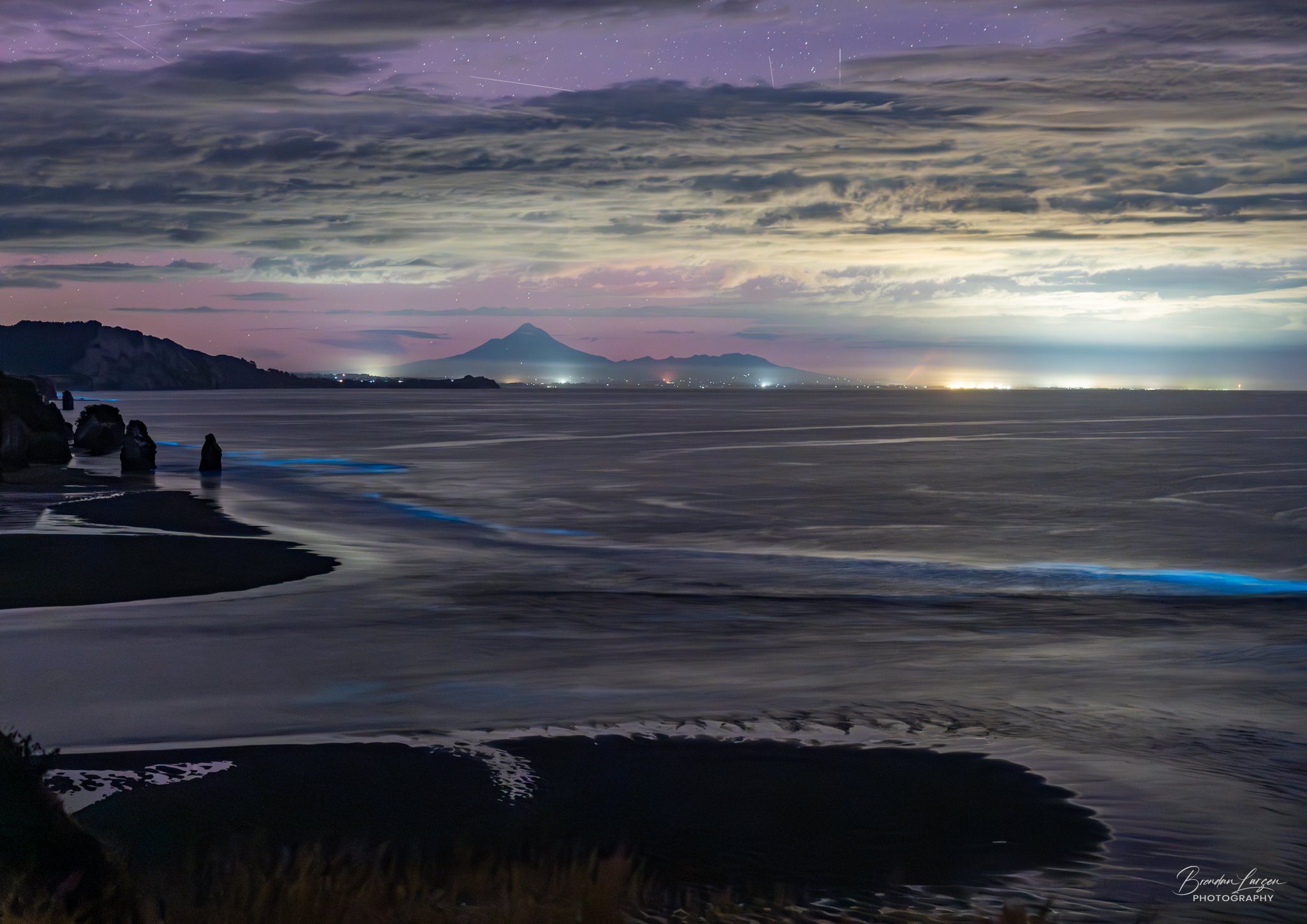 Nighttime view of a beach with rocks, water, and sand, overlooking an ocean. Distant mountains, including a prominent volcano, are visible under a dark, cloud-filled sky with faint stars.