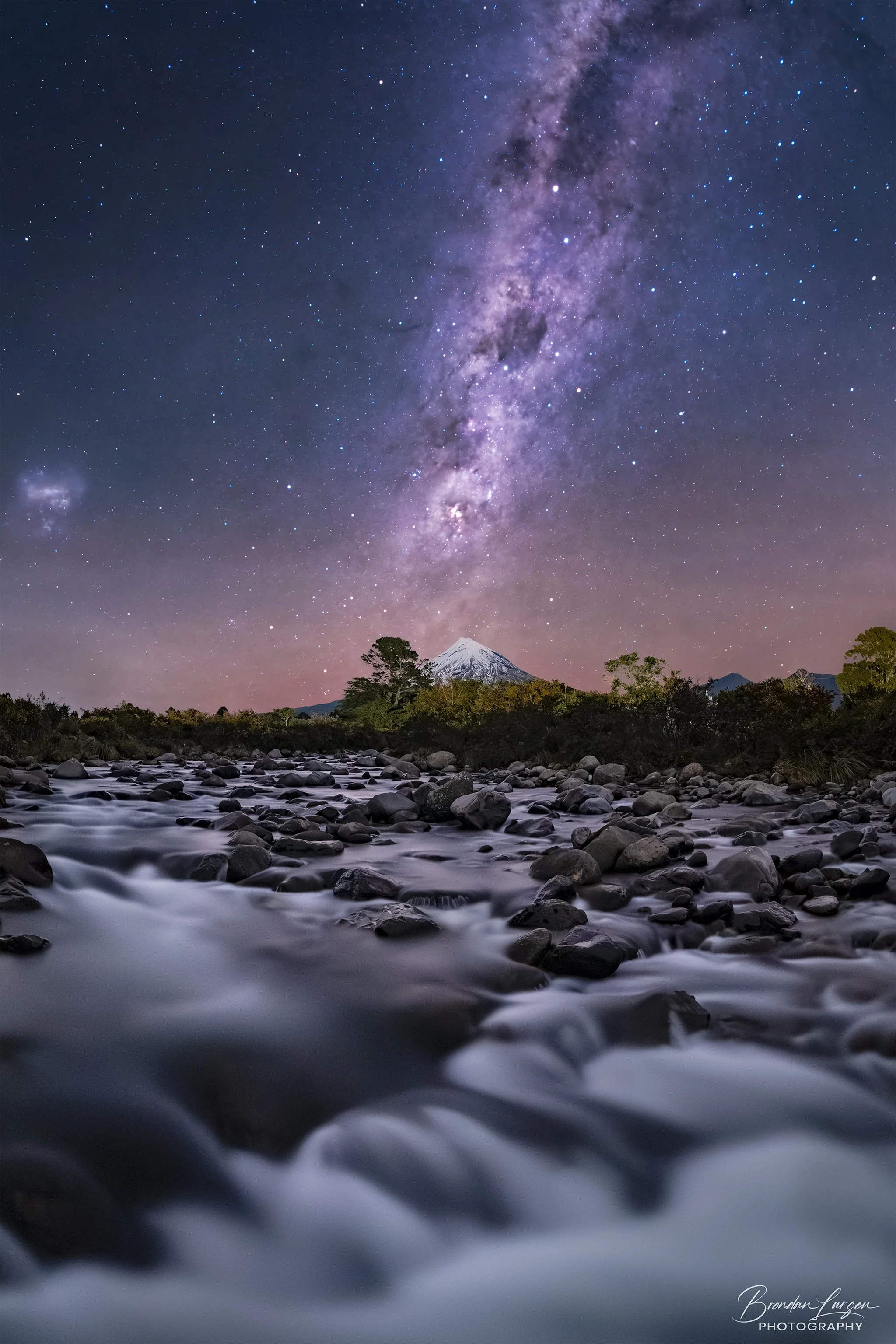 Night sky filled with stars and the Milky Way galaxy over a mountain with snow and a flowing river in the foreground.