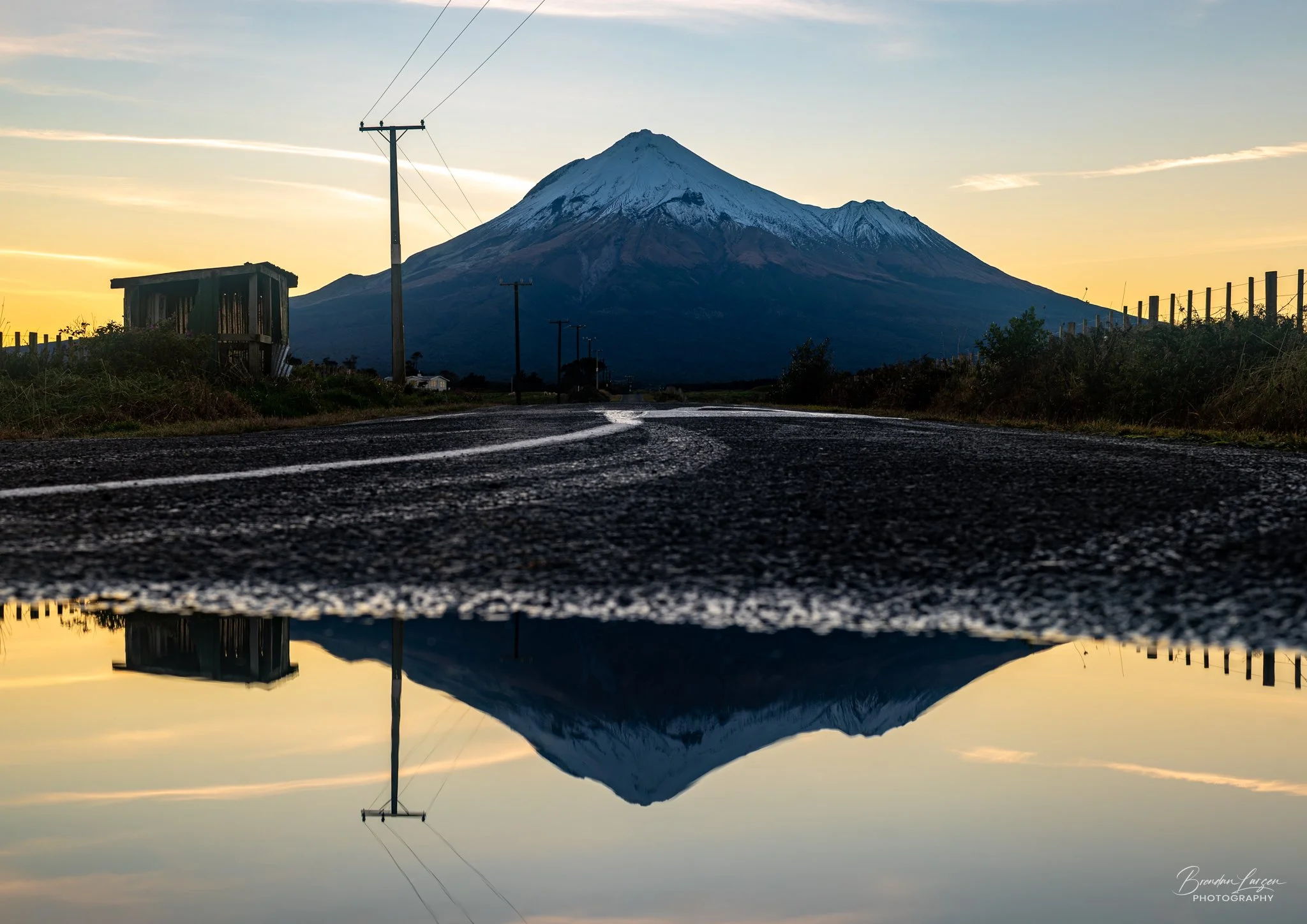 A scenic view of Mount Taranaki at sunset with a reflective puddle on the road in the foreground.