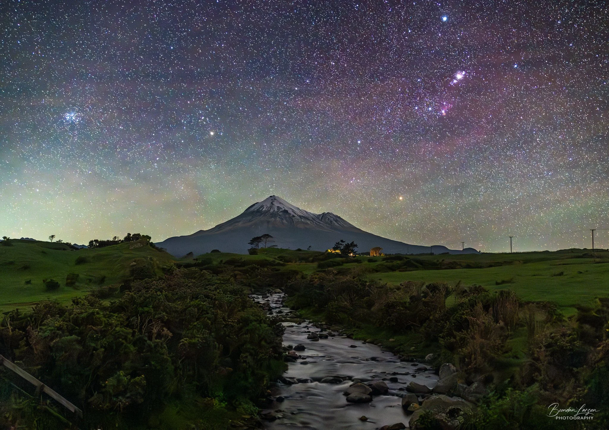 Nighttime landscape showing Mount Taranaki/Vulkan with snow on top, under a star-filled sky. A flowing stream runs through lush green fields with scattered trees.