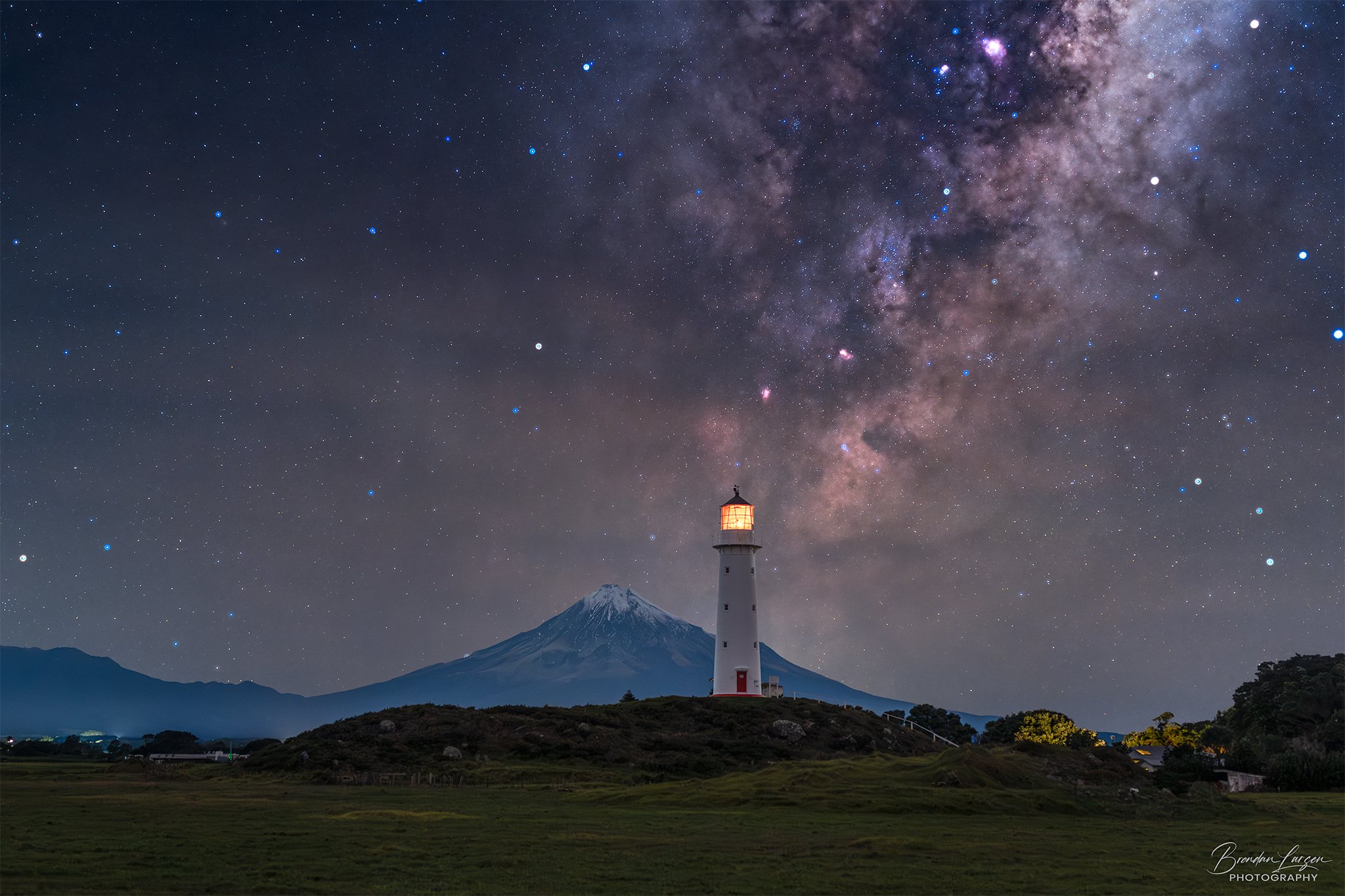 Nighttime landscape with a lighthouse on a grassy hill, Mount Taranaki in the background, and the Milky Way galaxy visible in the starry sky.