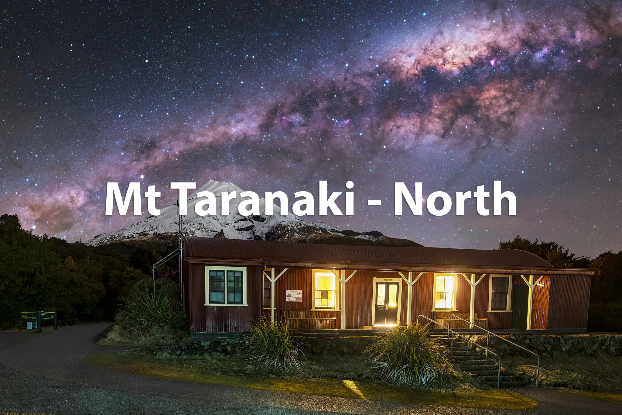 Nighttime view of a small building with lit windows against a backdrop of a snow-capped mountain and a star-filled sky with the Milky Way galaxy, labeled 'Mt Taranaki - North.'