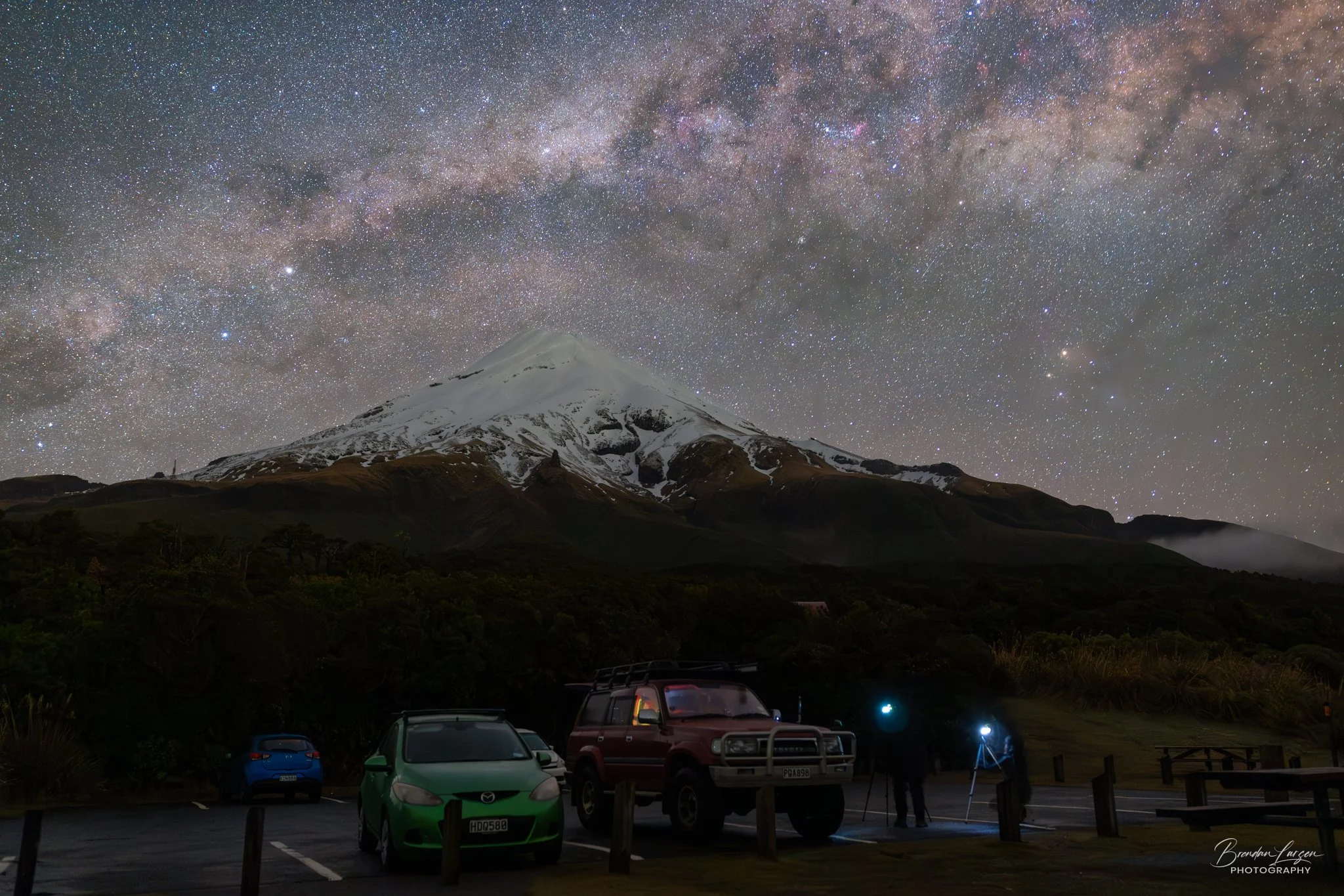 Night sky filled with countless stars and the Milky Way galaxy arching over a snow-capped mountain. In the foreground, several cars are parked, and two people are setting up telescopes to observe the stars.