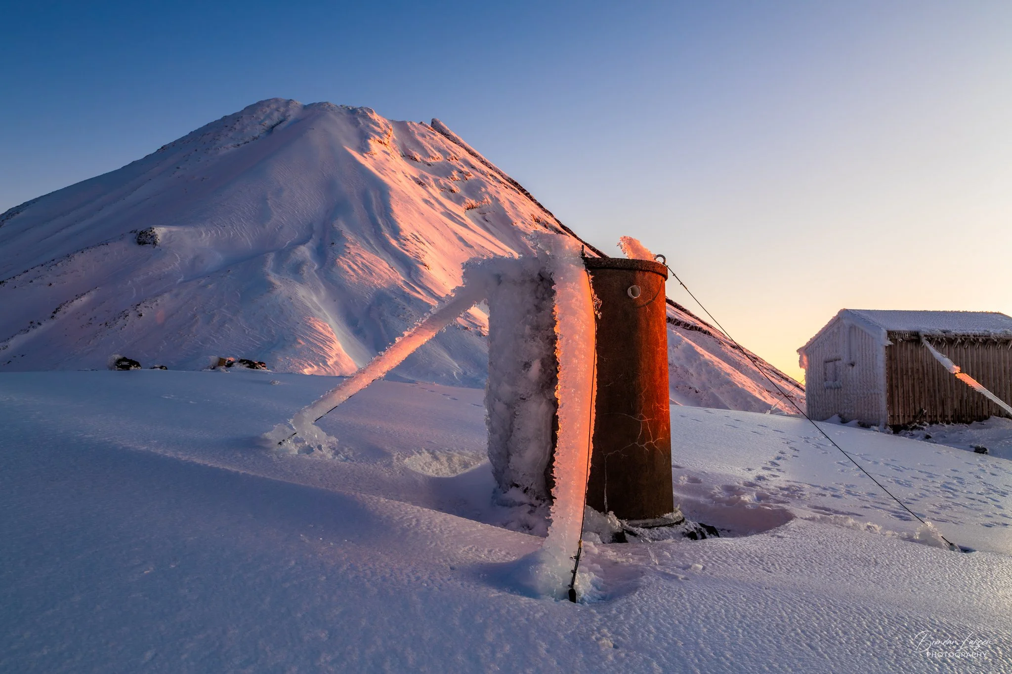 Snow-covered mountain with warm sunlight, snow-covered fence and small wooden building in the foreground.
