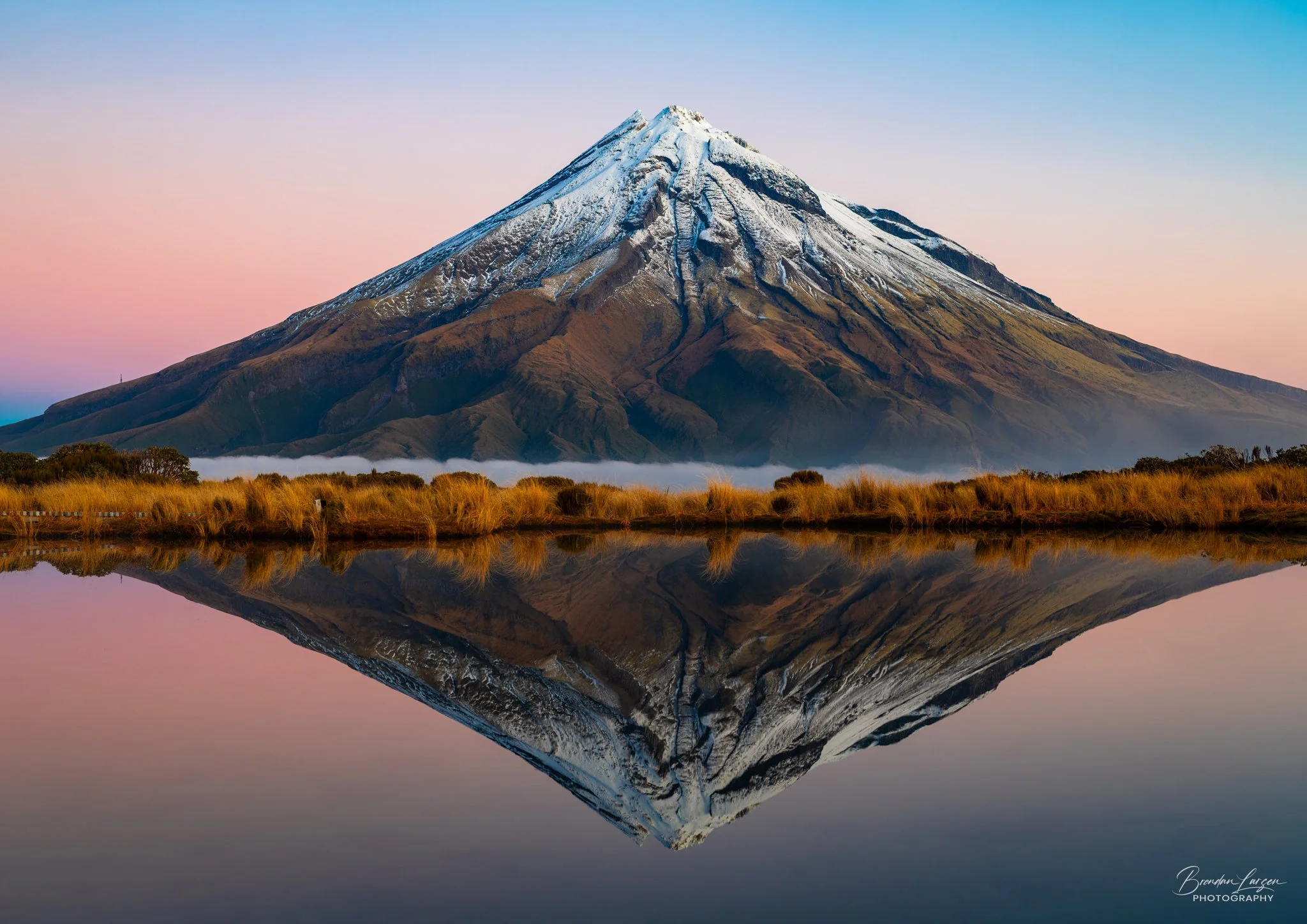 Snow-capped Mount Taranaki reflected in a calm body of water with grassy shoreline in the foreground, clear sky with soft pastel colors in the background.