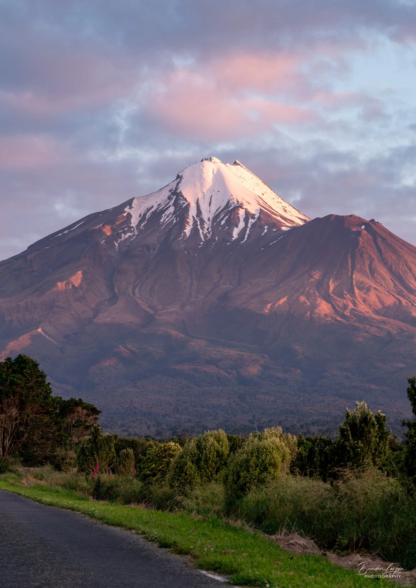 A snow-capped mountain, possibly Mount Taranaki, with a partly cloudy sky and greenery in the foreground.