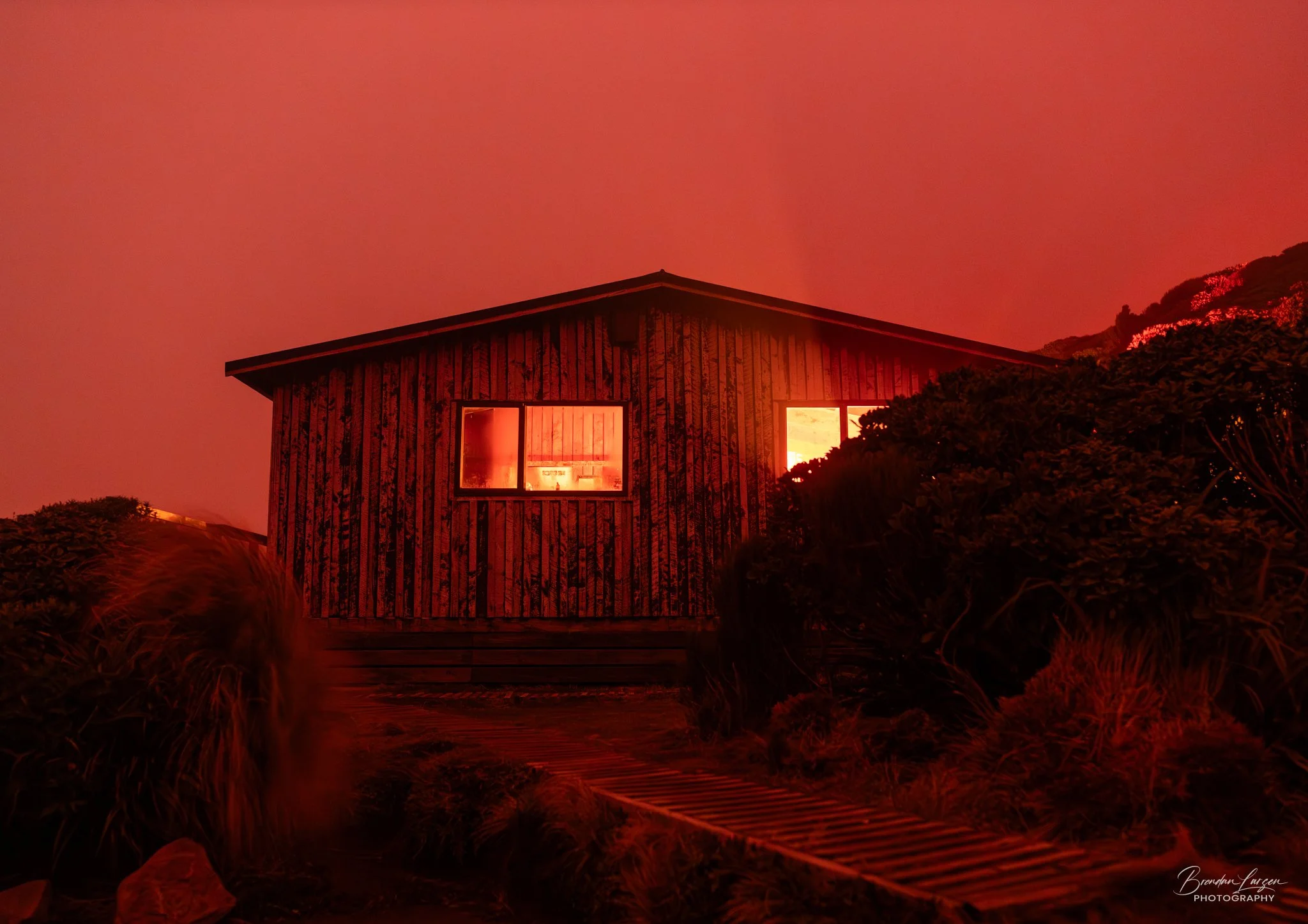 Photo of a wooden hut with lights on inside, surrounded by bushes and outdoor pathway under a red aurora sky.