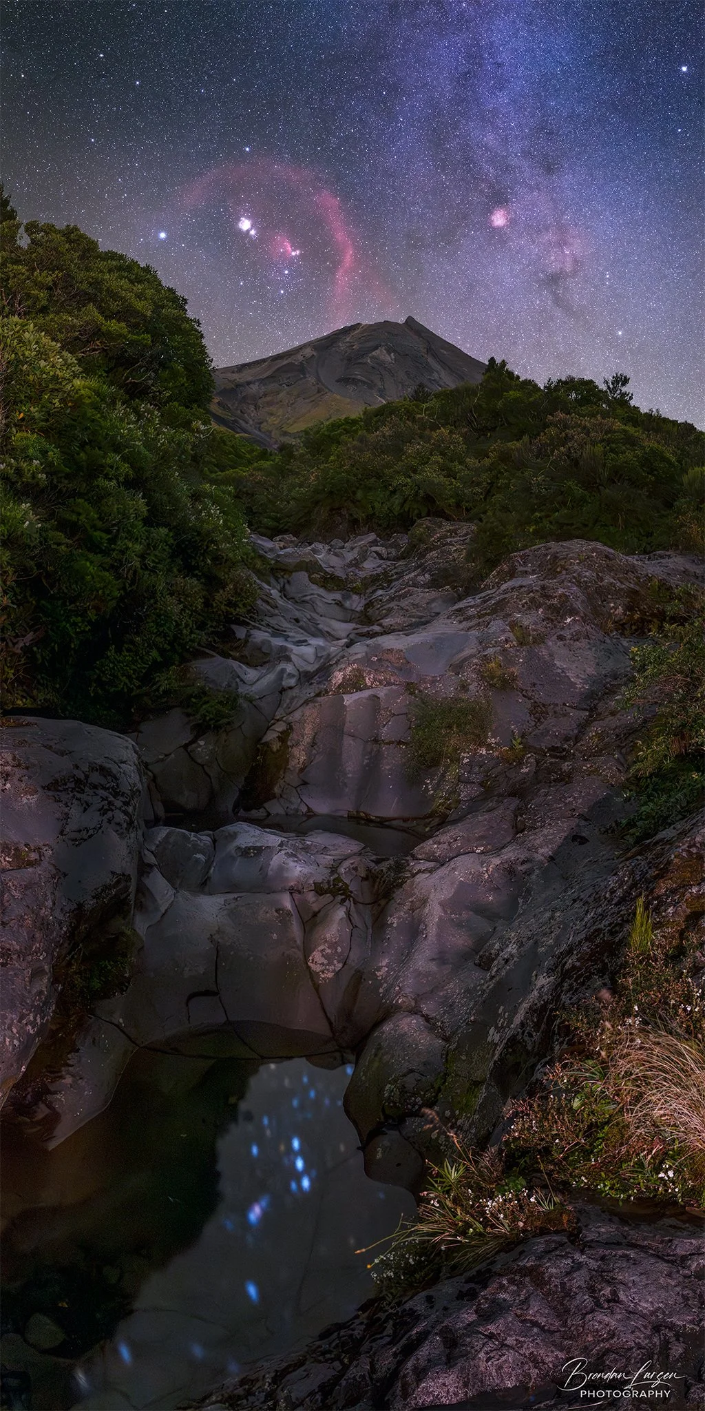 Nighttime landscape with starry sky, including the Milky Way, and a mountain in the background. Foreground features a rocky stream with a small pool reflecting the stars.