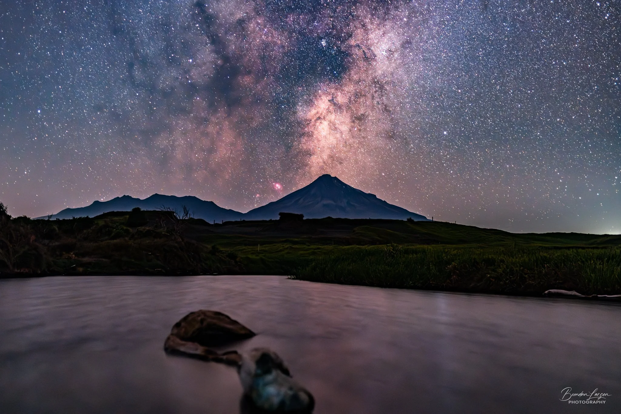 Nighttime landscape with a star-filled sky and the Milky Way galaxy over a silhouette of a mountain, with a body of water in the foreground.