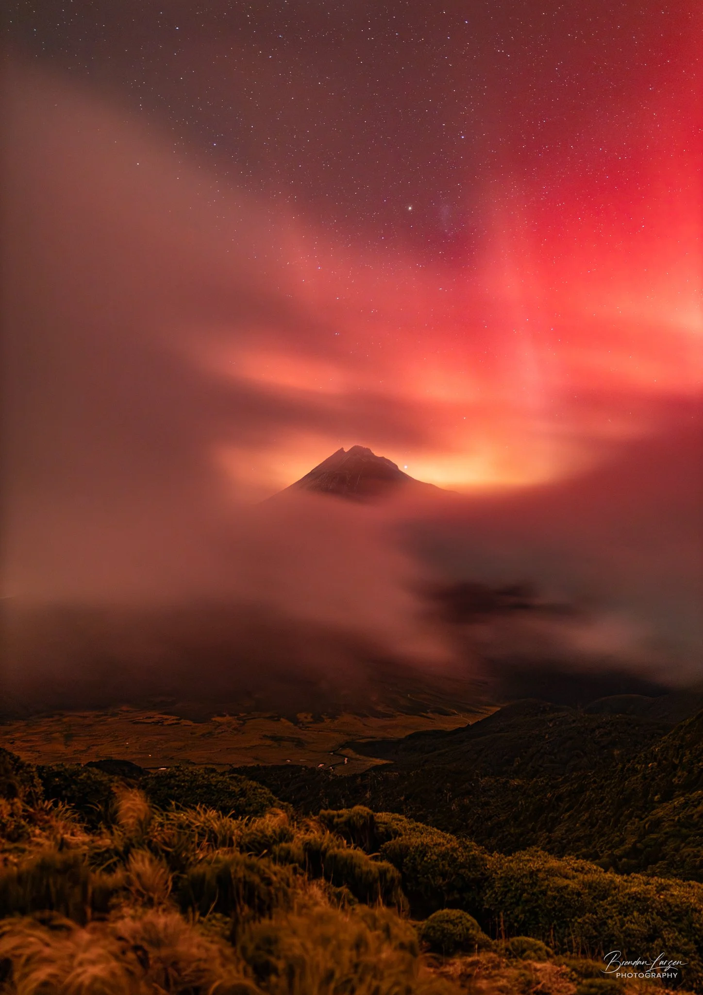 A mountain surrounded by clouds under a starry night sky with the aurora australis illuminating the sky in shades of red and pink.