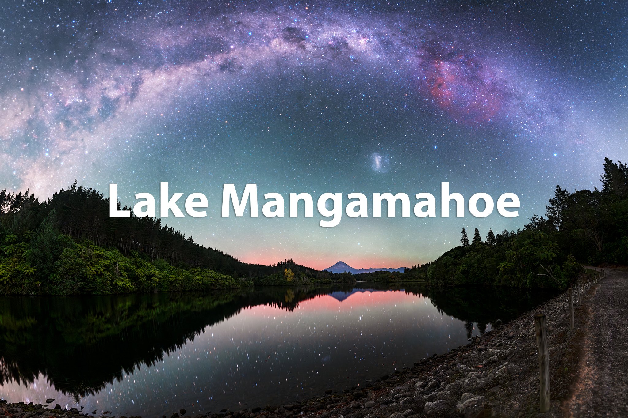 Nighttime view of Lake Manggapahoe with a starry sky, including the Milky Way galaxy, reflecting on the water, greenery on the edges, distant mountains, and a trail on the right.