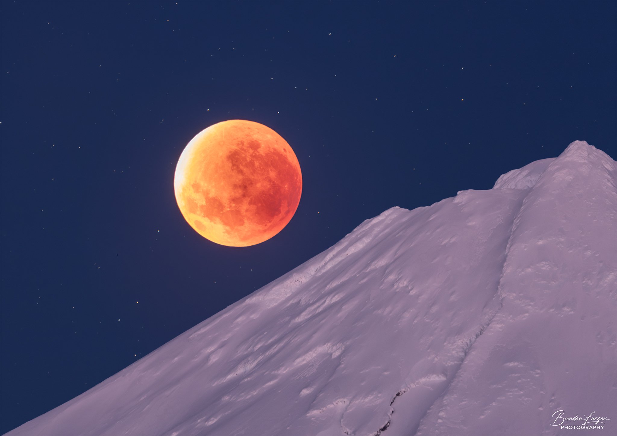 A blood moon during a lunar eclipse over a snow-covered mountain at night.