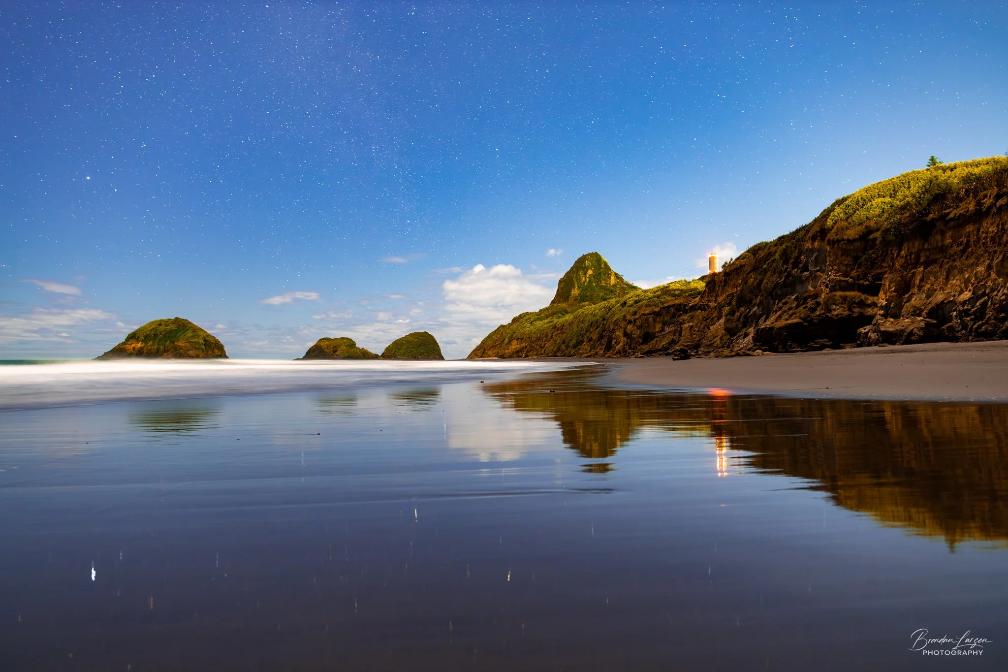 Nighttime view of a rocky beach with large green cliffs and island formations, stars visible in the sky, and a lighthouse on one of the cliffs illuminated and reflected in the water.