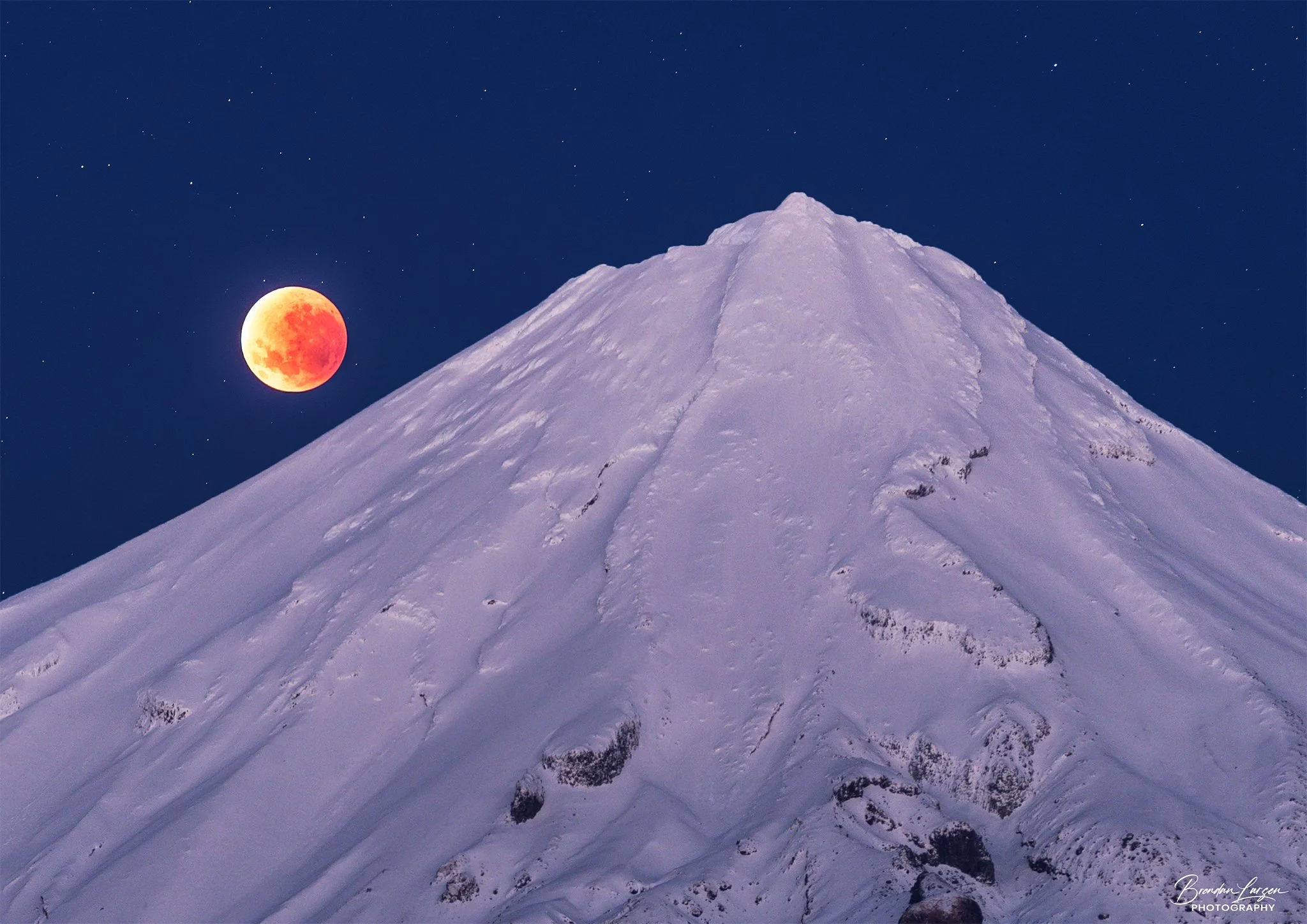 A snow-covered mountain peak under a dark night sky with a blood moon and stars.
