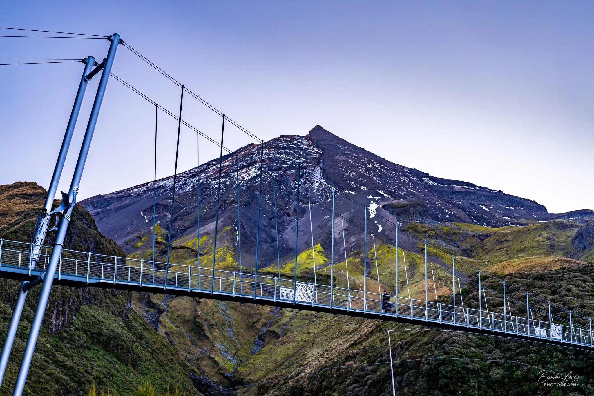 Suspension bridge spanning a lush green valley with a mountain in the background, snow patches on the mountain, clear blue sky.