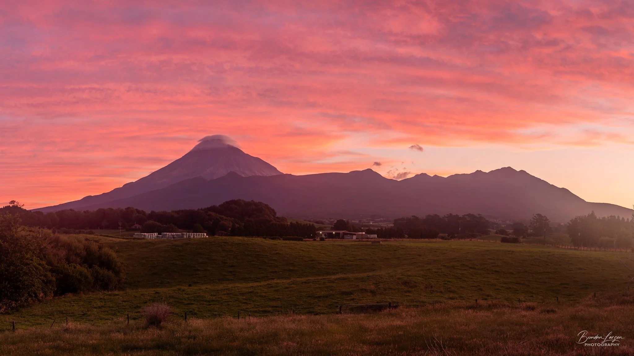 A scenic view of a mountain with a volcano-like peak at sunset, pink and orange clouds in the sky, green grass and trees in the foreground.