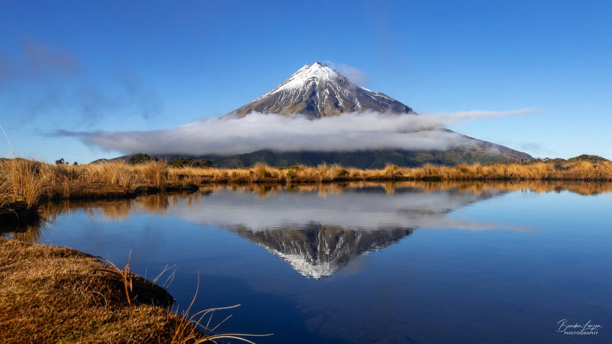 A snow-capped mountain reflected in a lake with grassy banks under a clear blue sky.