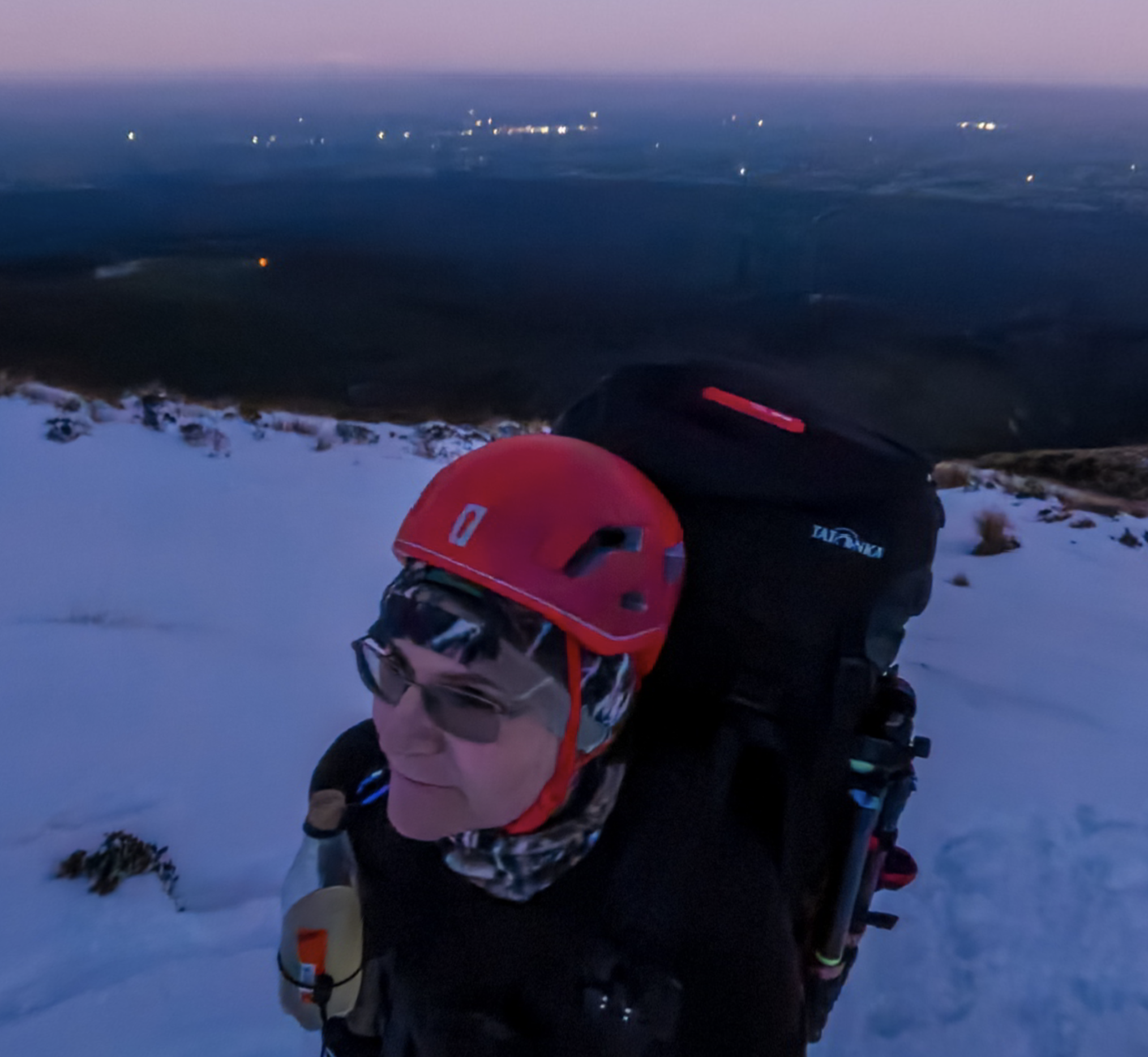 A person wearing a red helmet, sunglasses, and a scarf, carrying a backpack, standing on snowy terrain during dusk or dawn, with a view of a landscape and city lights in the distance.