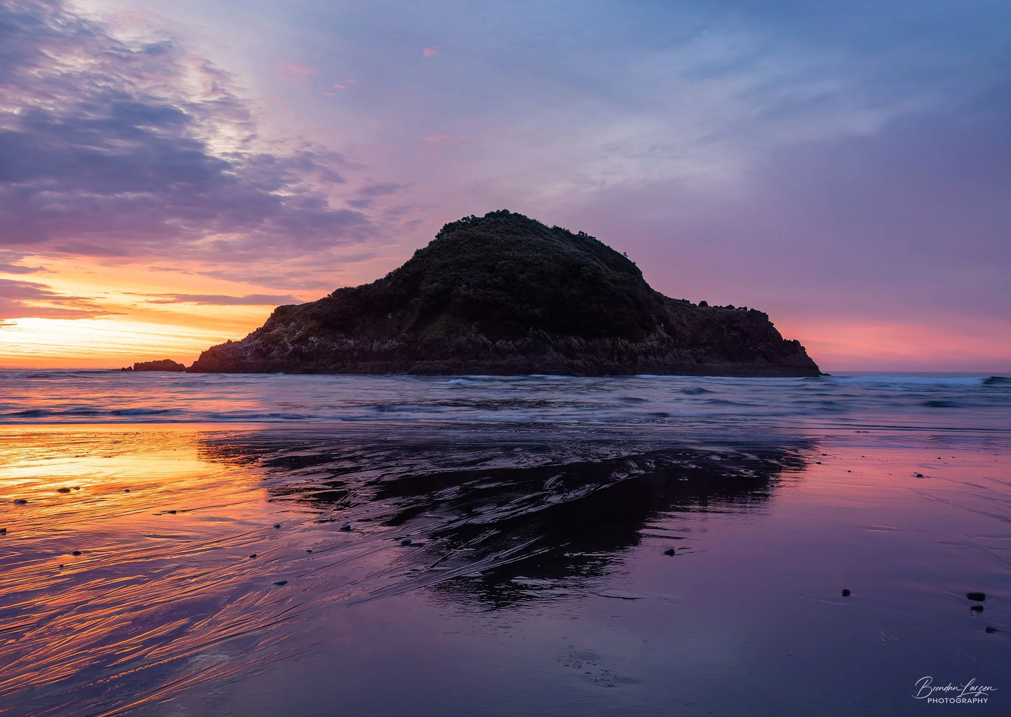 A scenic view of a rocky island during sunset with colorful sky and reflections on the wet sand in the foreground.