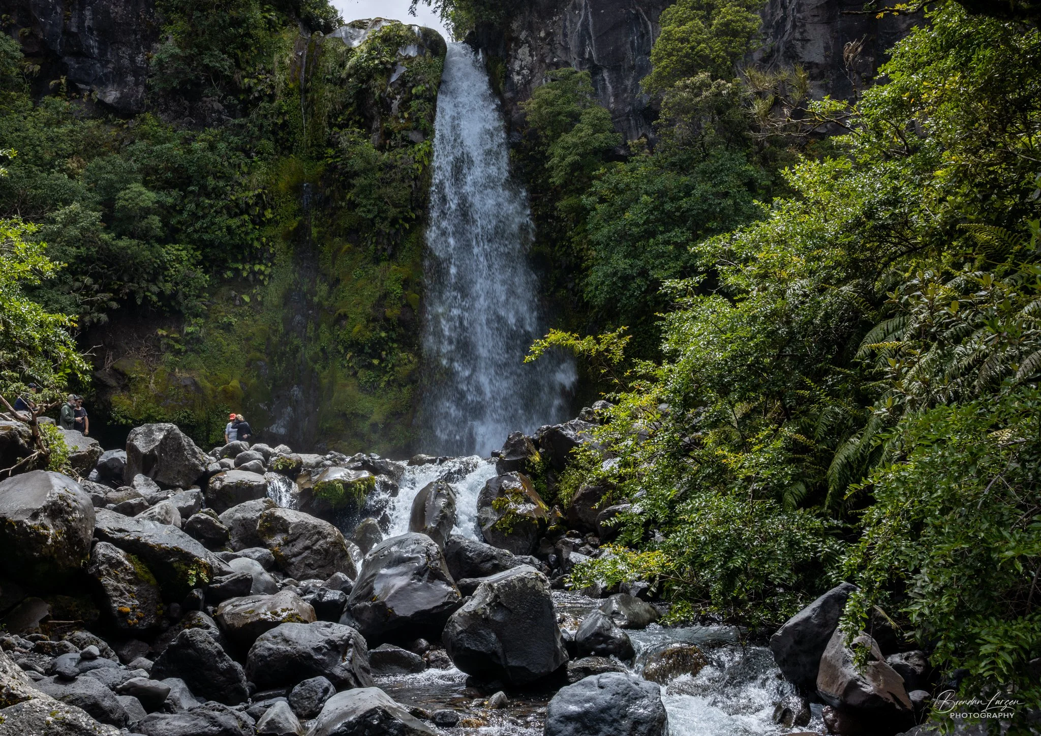 A waterfall cascading down a rocky cliff surrounded by lush green foliage, with a few people sitting on rocks near the water.