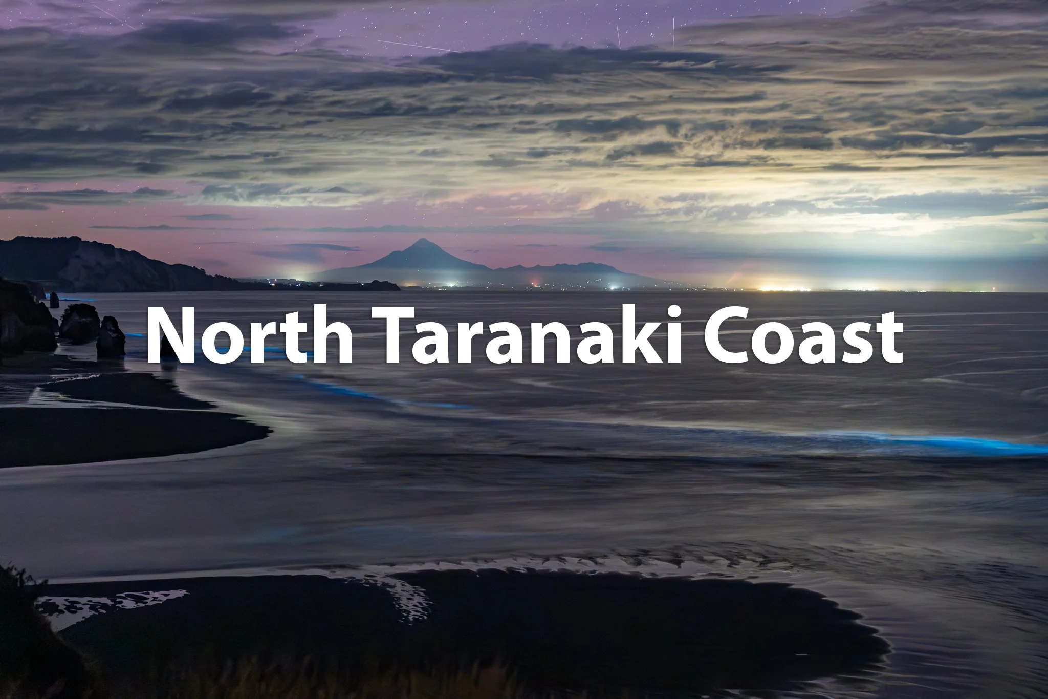 Nighttime view of the North Taranaki Coast with dark clouds, mountain in the distance, and the ocean in the foreground.