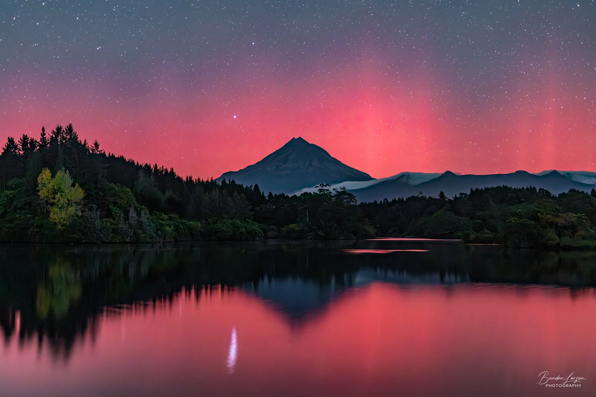 A mountain with a peak in the distance under a starry sky with a pink sunset, reflected in a calm body of water surrounded by trees.