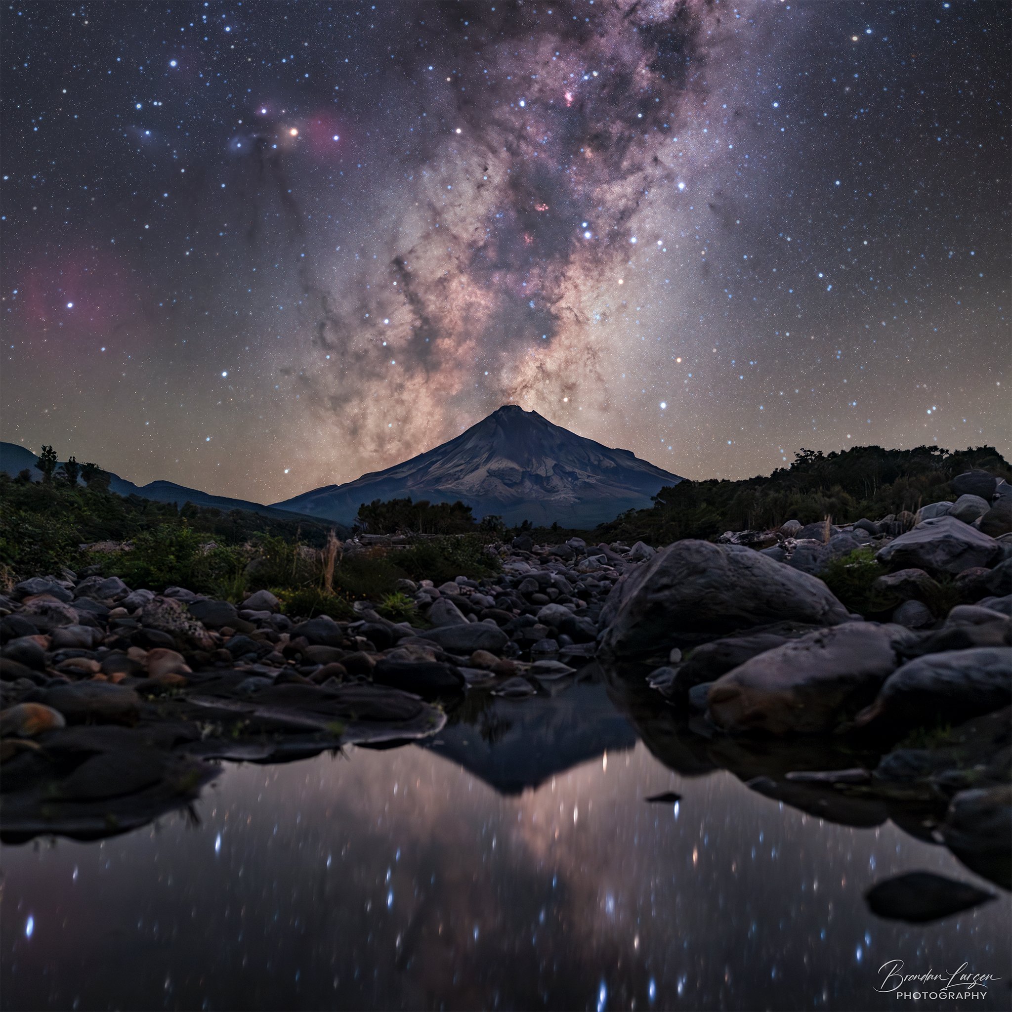 Night sky with stars and the Milky Way galaxy above a mountain, with a rocky foreground and reflection in a small pool of water.