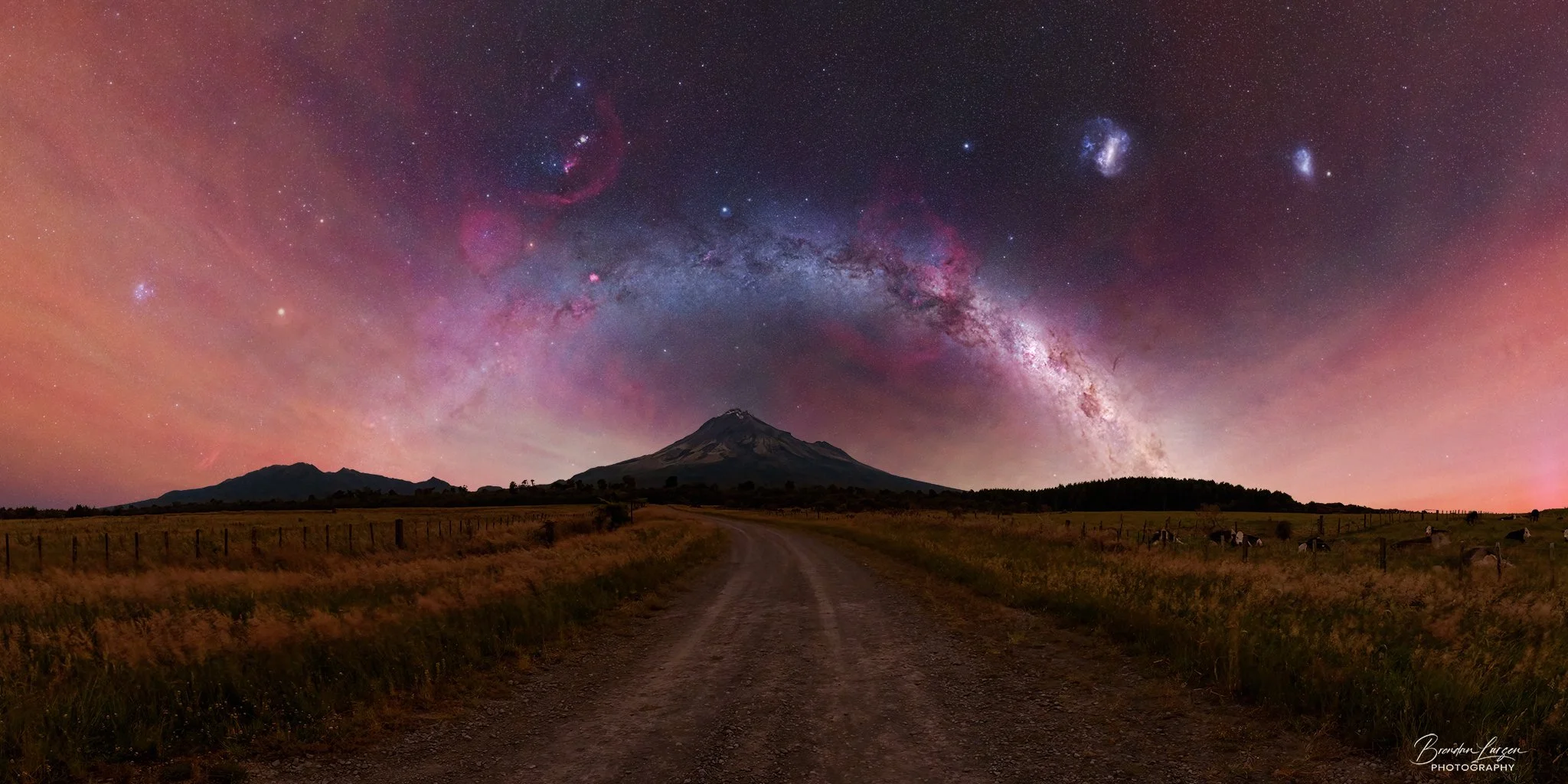 Night sky over a mountain with visible Milky Way galaxy and stars, a dirt road leading to the mountain, fields with grazing cows in foreground.