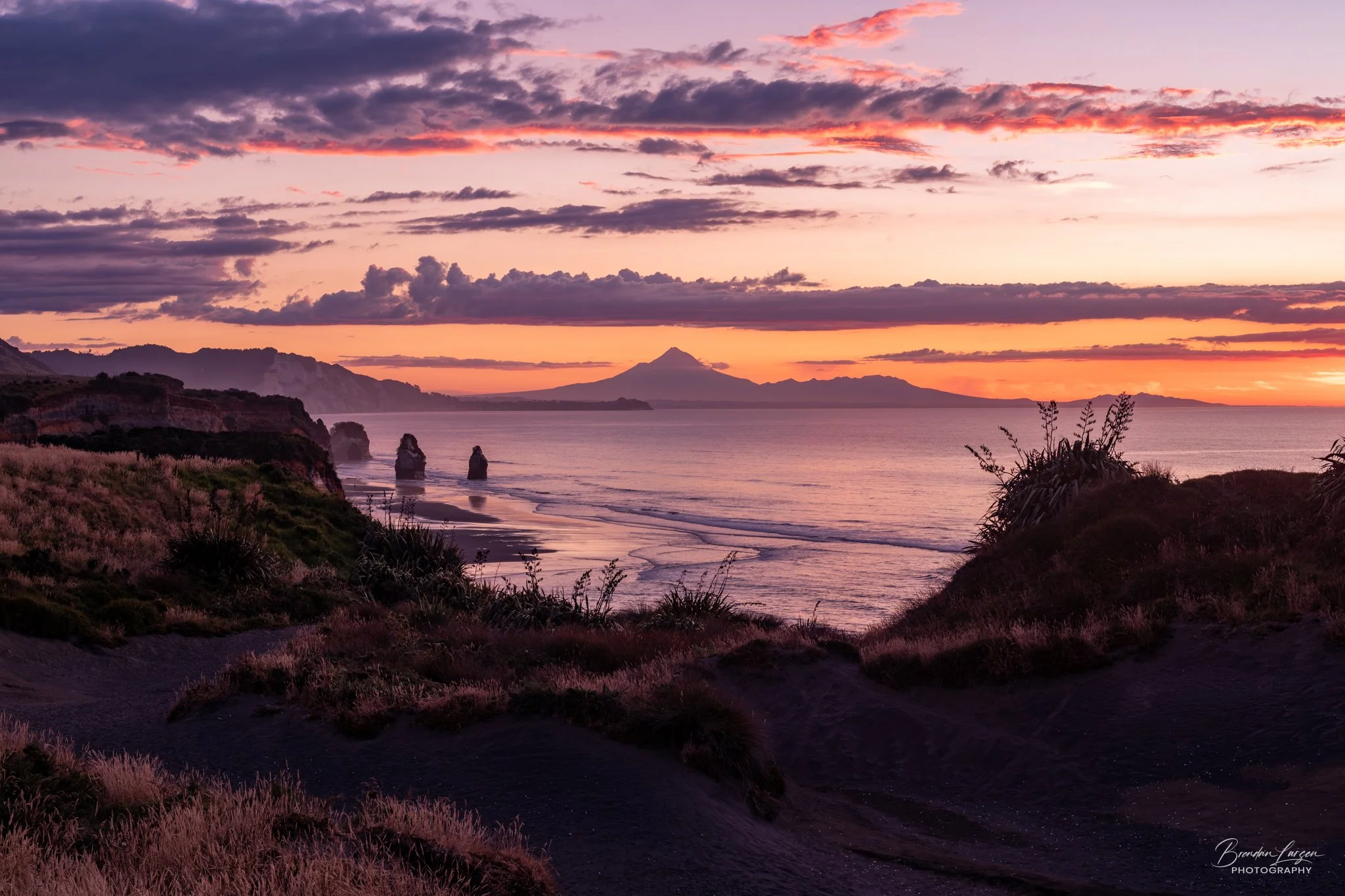 Sunset over a beach with colored clouds, distant mountains, and ocean waves, with grassy dunes in the foreground.