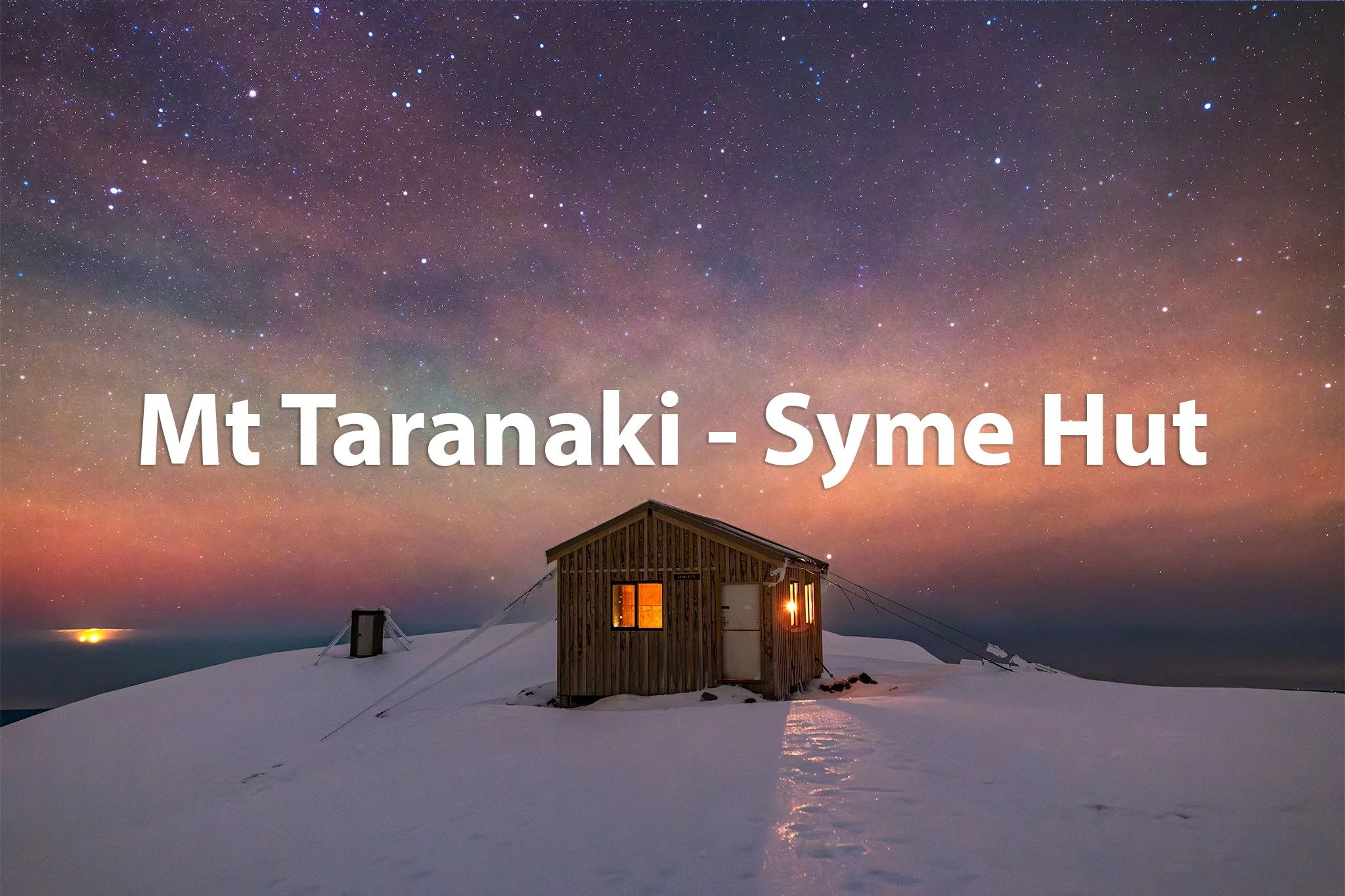 Snow-covered landscape with a small wooden hut emitting warm light under a starry night sky with the Milky Way, located at Mount Taranaki, Syme Hut.