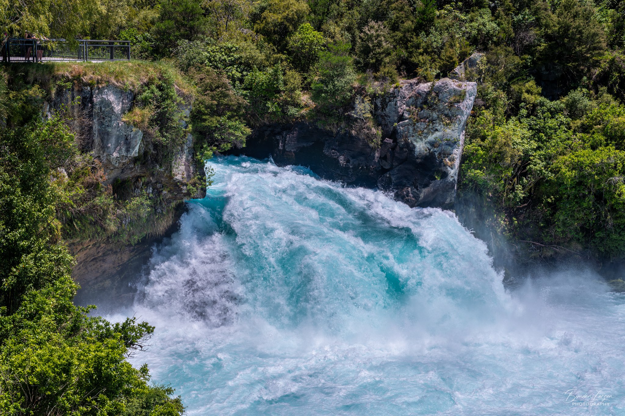 Fast-flowing river with turquoise water rushing through a rocky waterway surrounded by lush green trees and bushes.