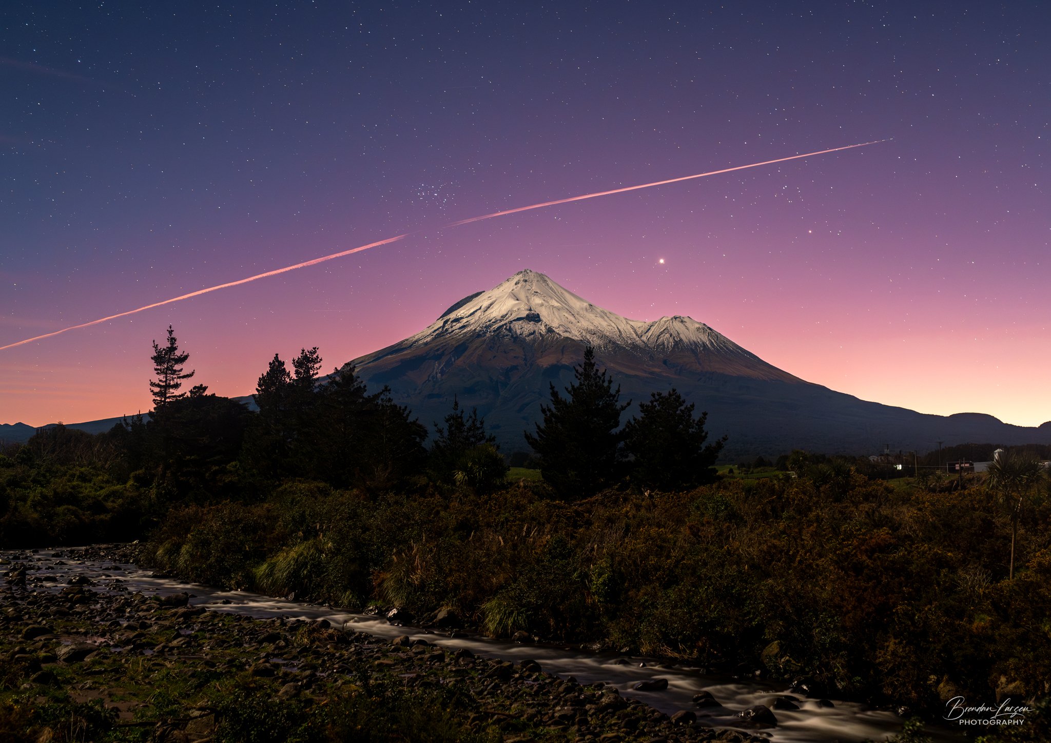 A snow-capped mountain, likely Mount Taranaki, during twilight with a colorful sky. Stars and a streak of pink and orange light are visible in the sky. Trees and a rocky stream are in the foreground.